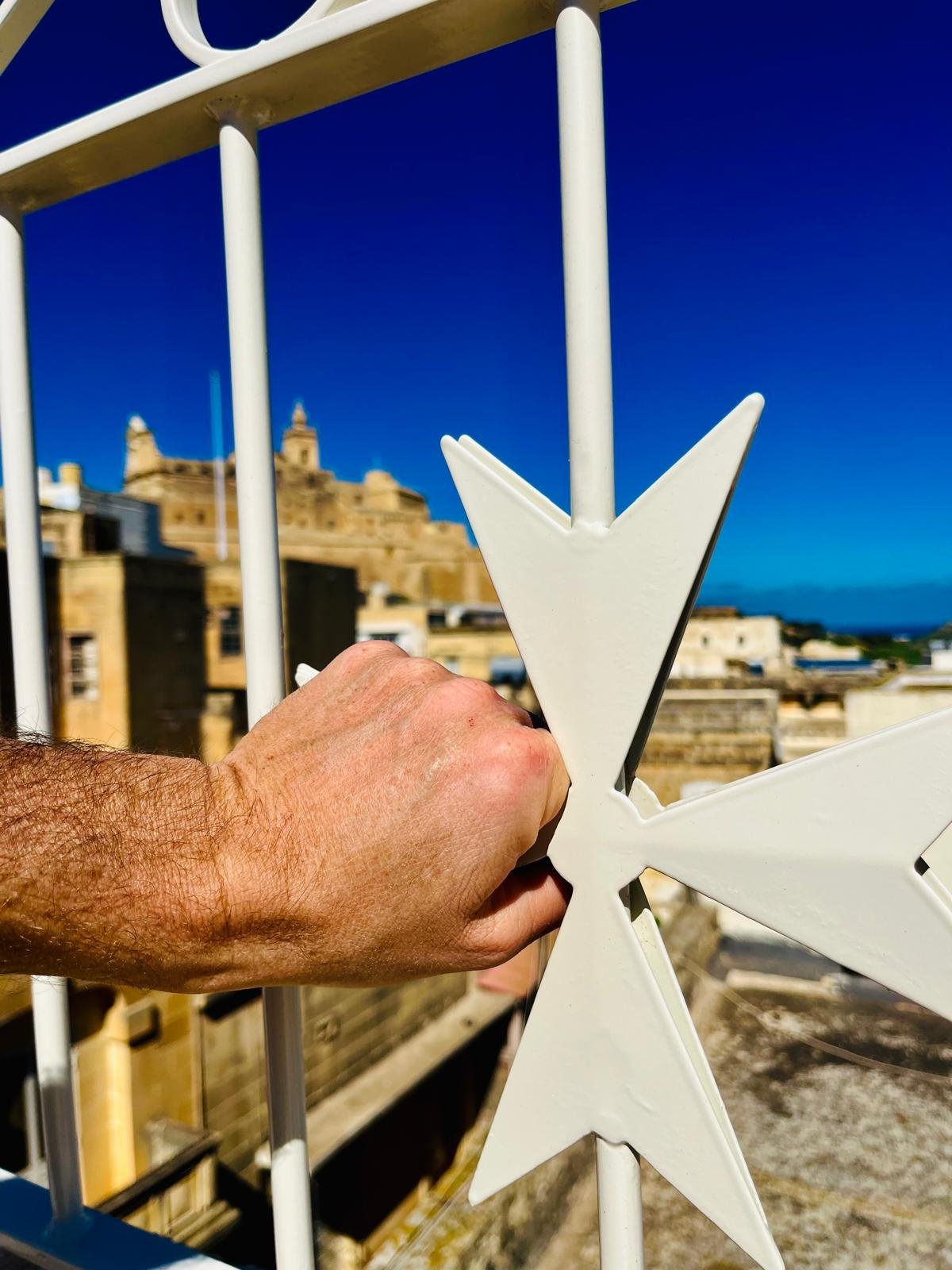 A person's hand gripping a white star-shaped ornament on a metal fence, with a historical building and blue sky in the background.