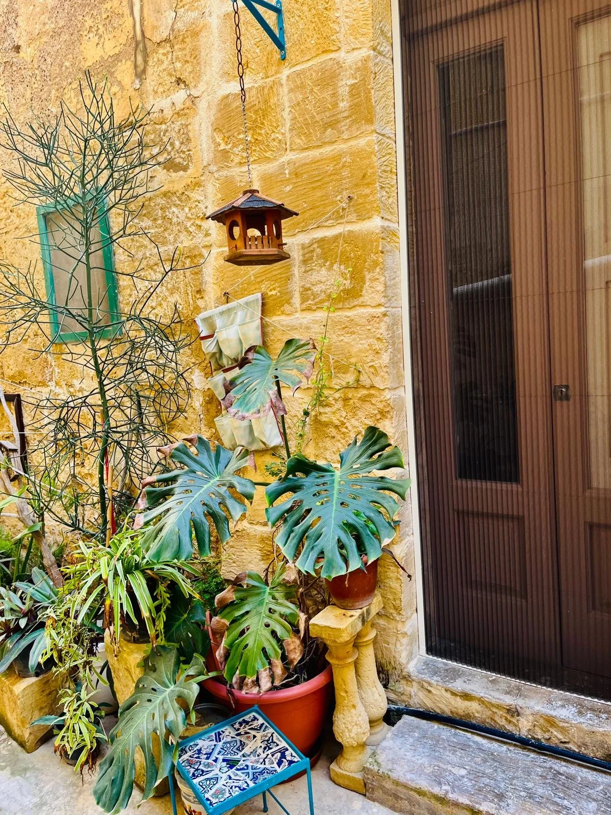 Outdoor corner with yellow stone wall, potted plants, a birdhouse, a small decorative table, and a sliding glass door.