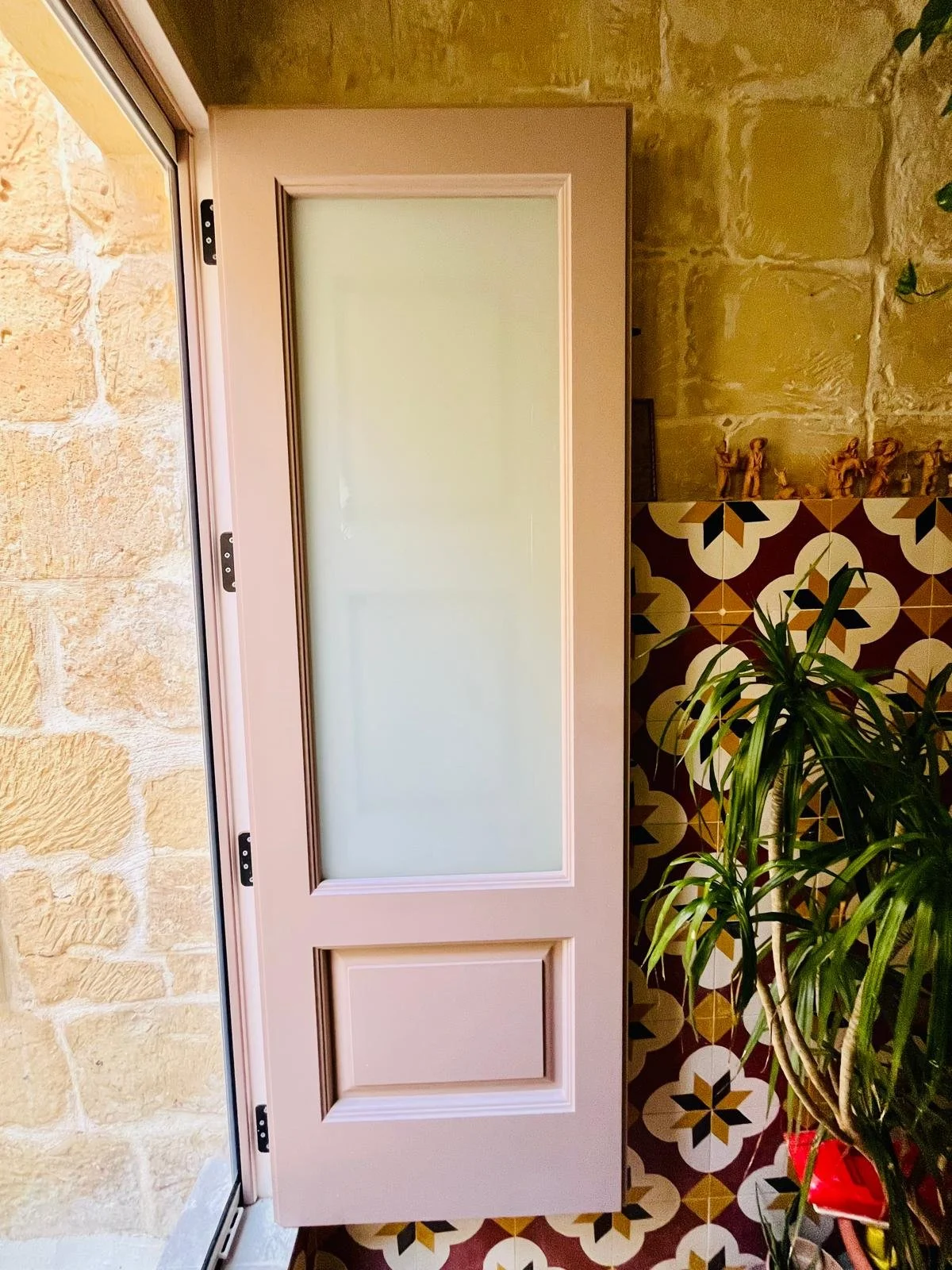 Pink door with frosted glass panel, partially open, next to a brick wall, with a patterned tile floor and a green potted plant nearby.