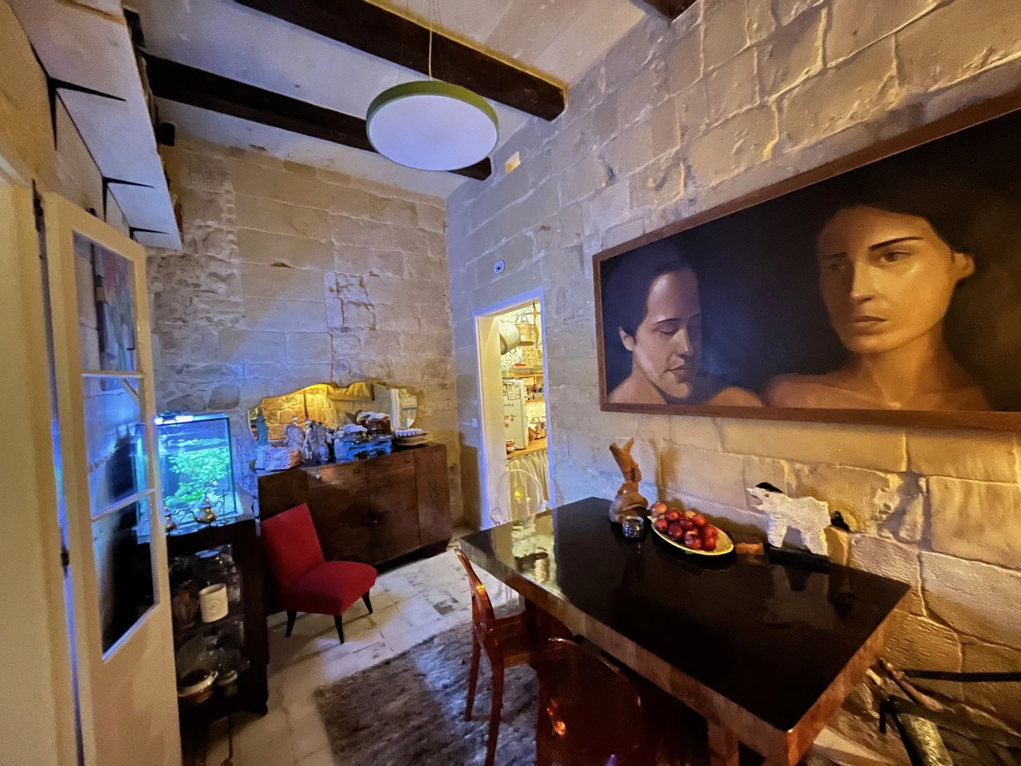A dining room with stone walls, a black table with a bowl of red apples, a red chair, a wooden sideboard with a mirror, a fish tank, and a large painting of two women on the wall.