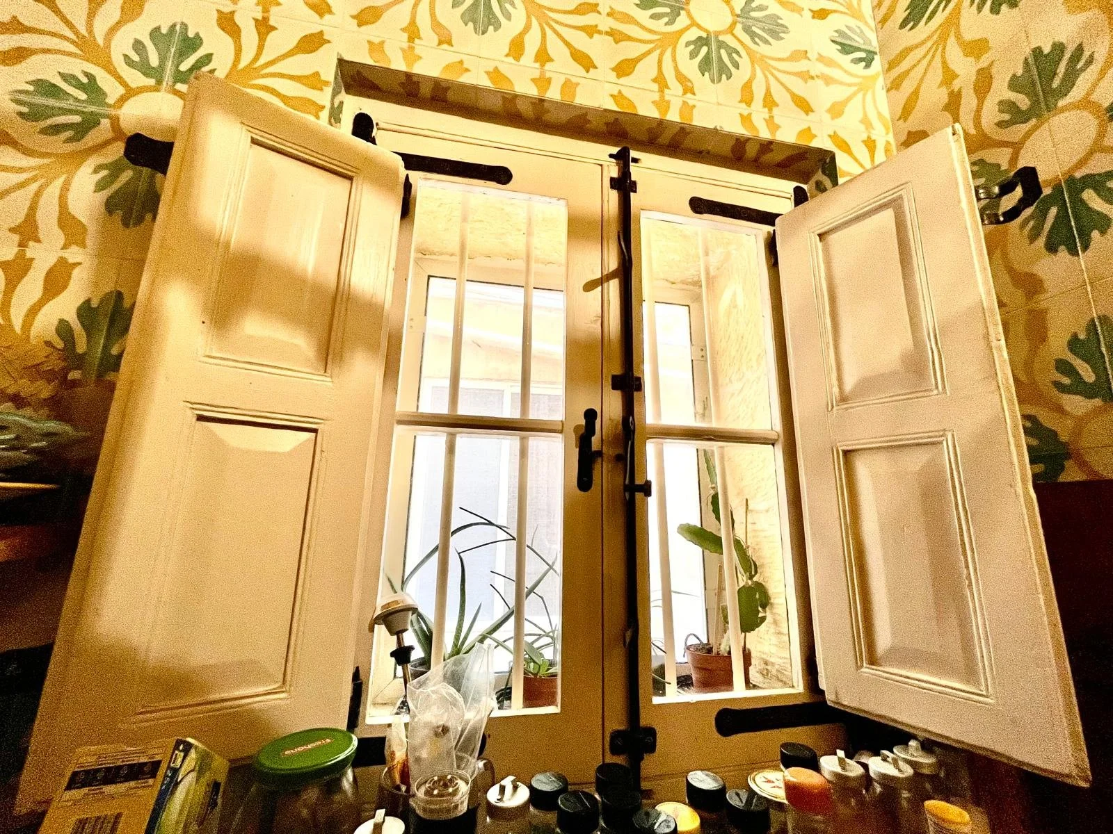 Open window with white shutters and black hinges, metal bars, and a latch, overlooking a patio with potted plants. The wall features a yellow and green wallpaper pattern.
