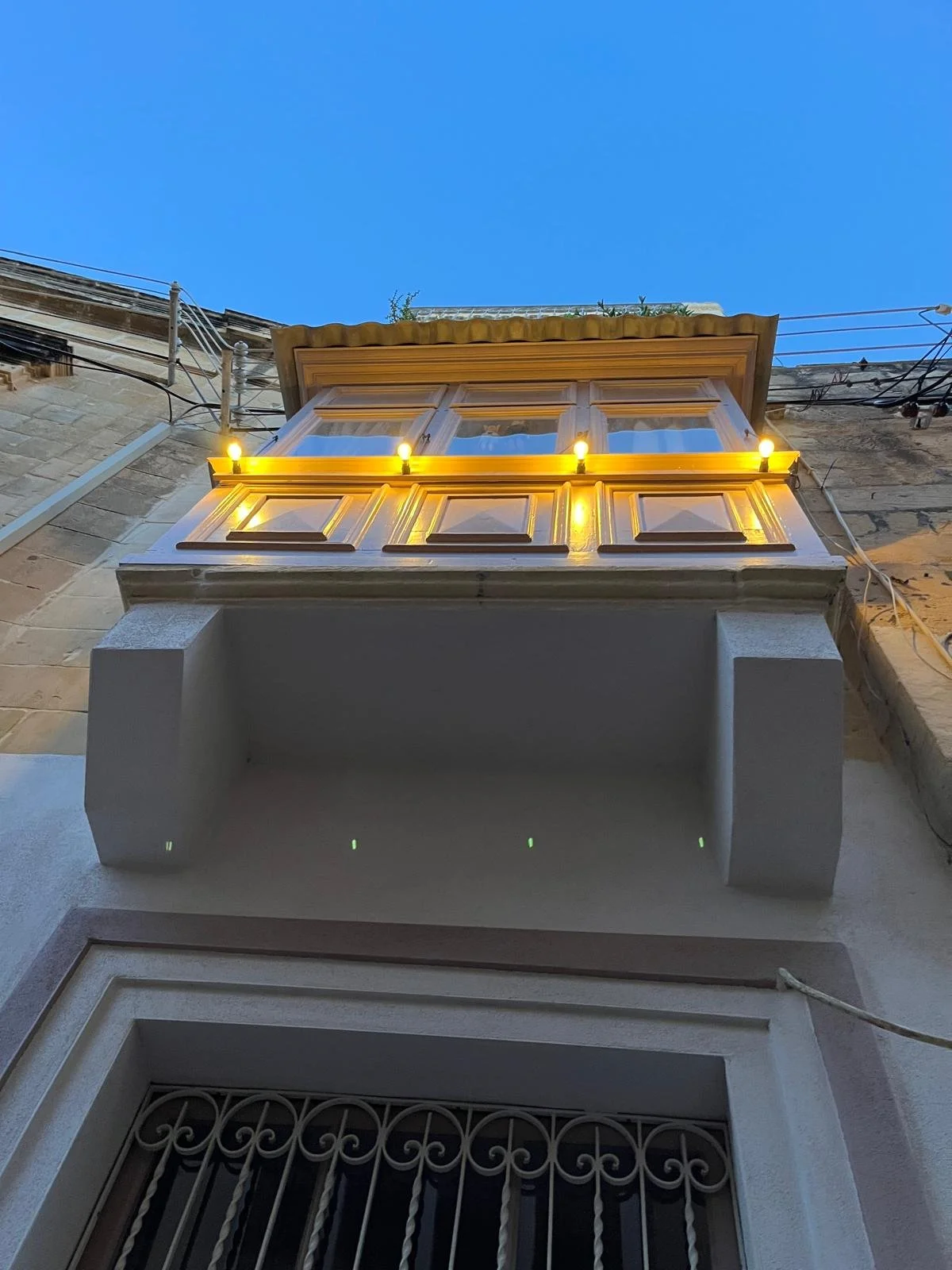 View of a building facade from an upward angle showing a balcony with wooden panels, a set of yellow string lights, and a window with metal bars, against a blue evening sky.