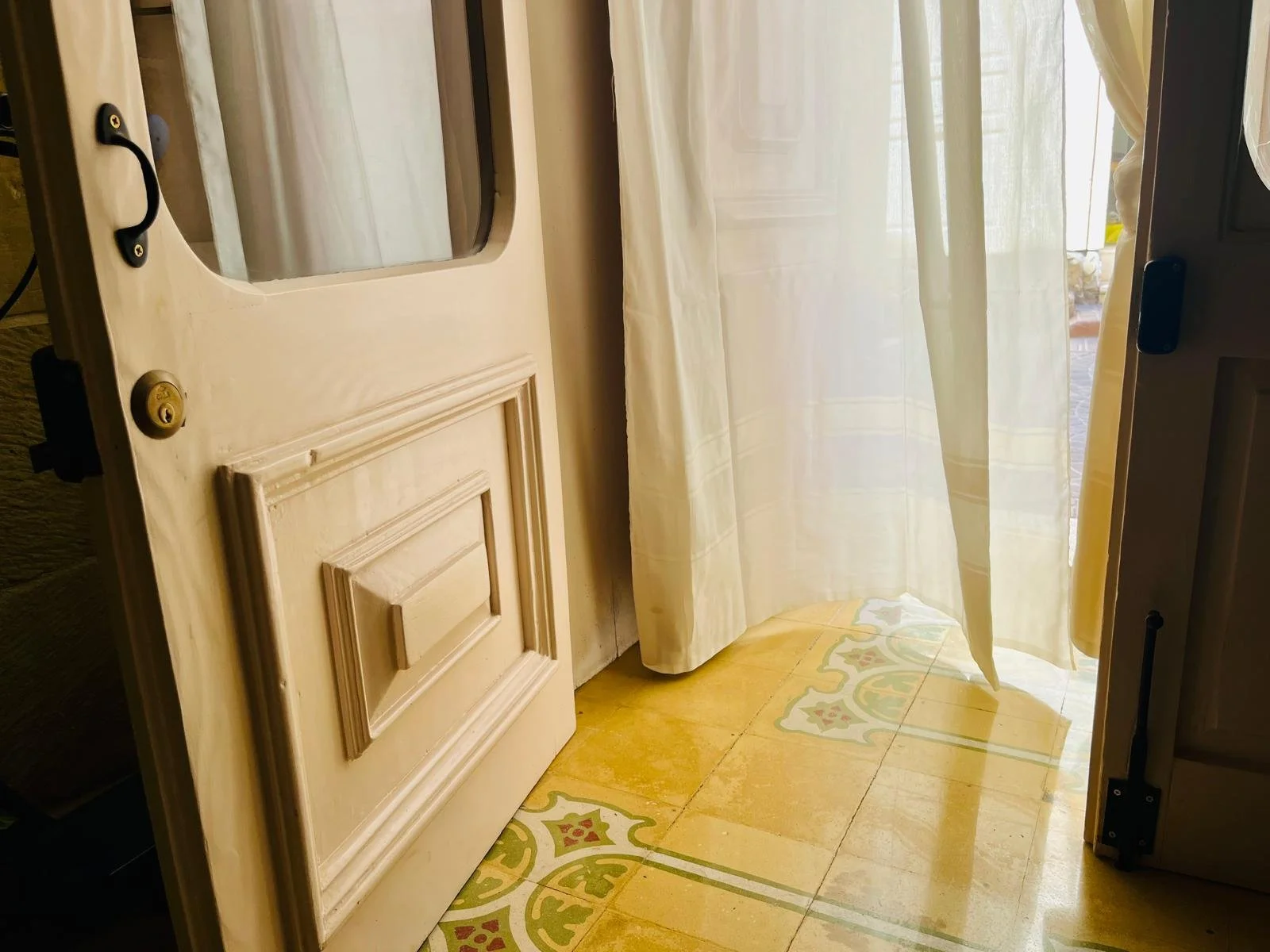 An open door revealing a room with a yellow tiled floor, patterned with green and red accents, and a sheer curtain leading outside to a bright area.