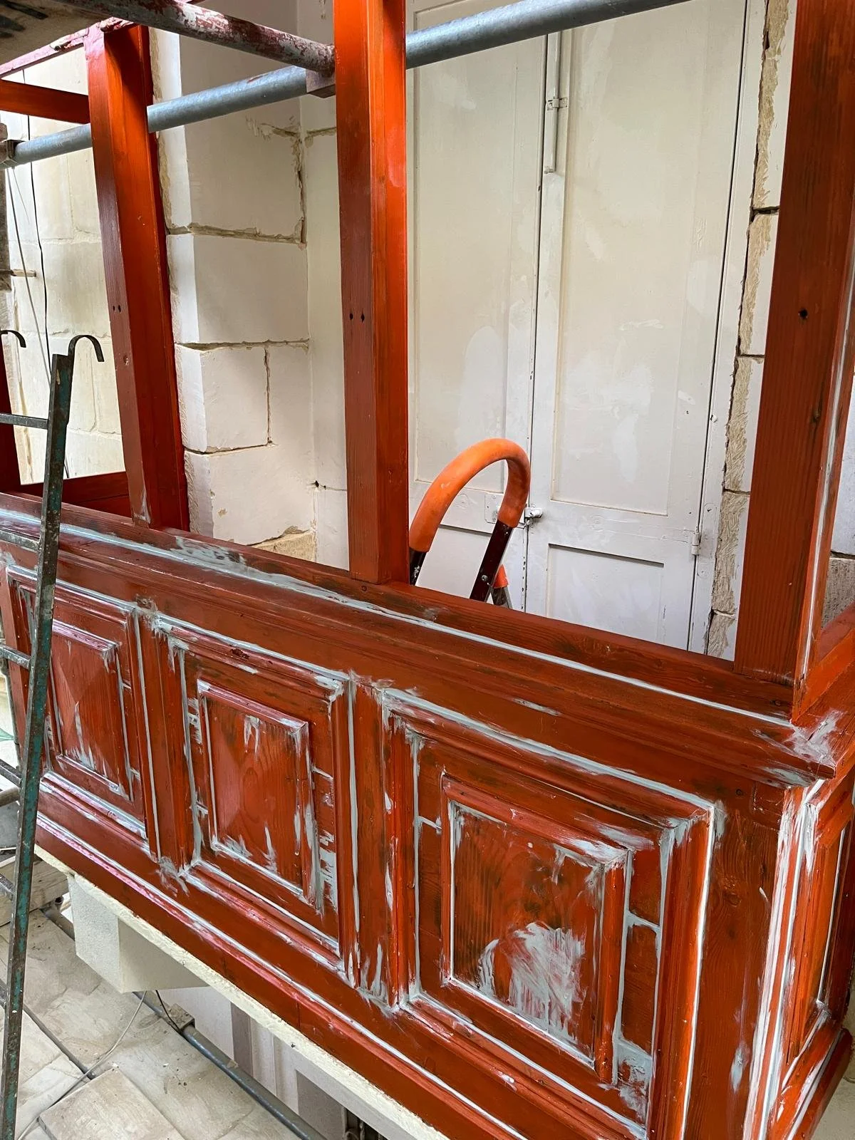 Close-up of a wooden cabinet or furniture piece in the process of being painted or restored, with visible paint and primer patches, and a stepladder nearby.
