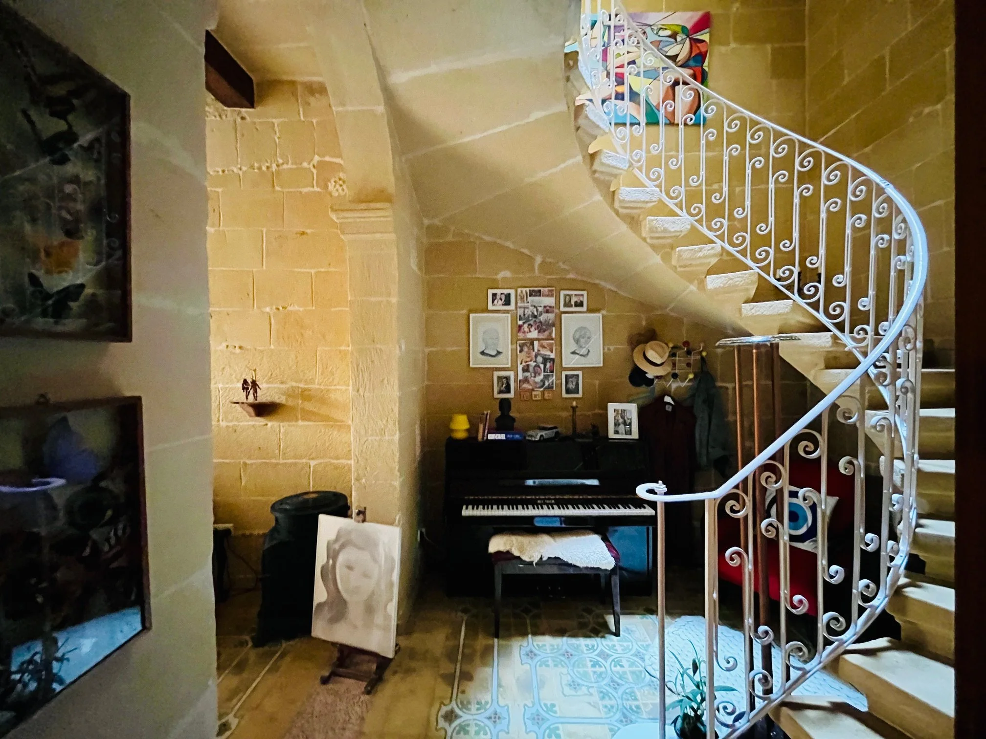 Interior view of a cozy room with a spiral staircase, a black piano, framed photographs on the wall, a portrait on an easel, and various decorative items.