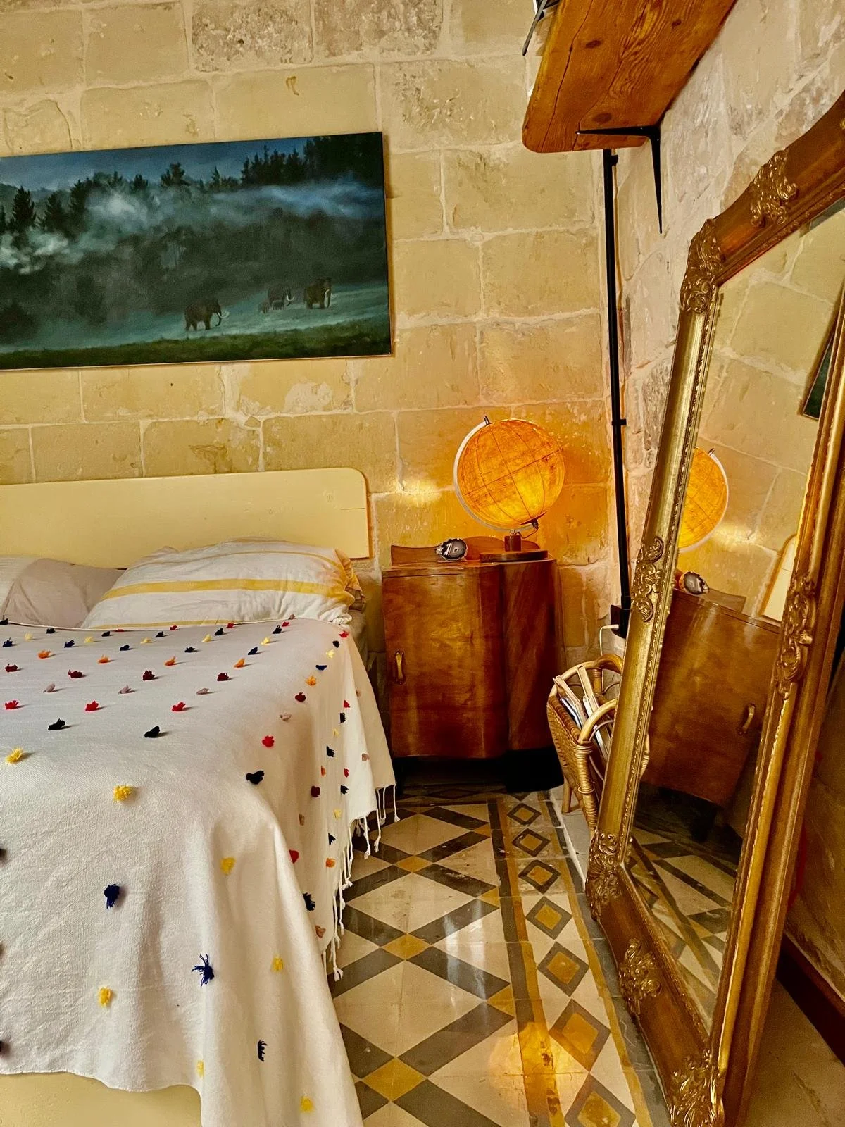 A bedroom with a bed covered in a white blanket decorated with small colorful pom-poms, next to a wooden nightstand with a globe-shaped lamp, a framed mirror leaning against the wall, a painting of elephants in a misty forest, and a patterned tile fl