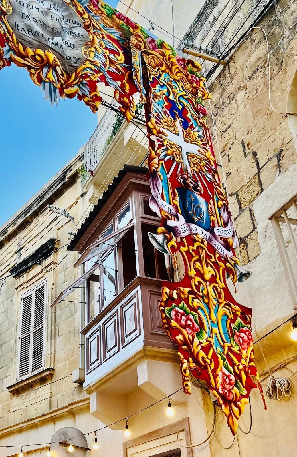 Colorful decorative hanging banner with a coat of arms, featuring a cross, floral patterns, and Latin inscriptions, hanging outside a building with stone walls and windows, with string lights visible below. Clear blue sky in the background.