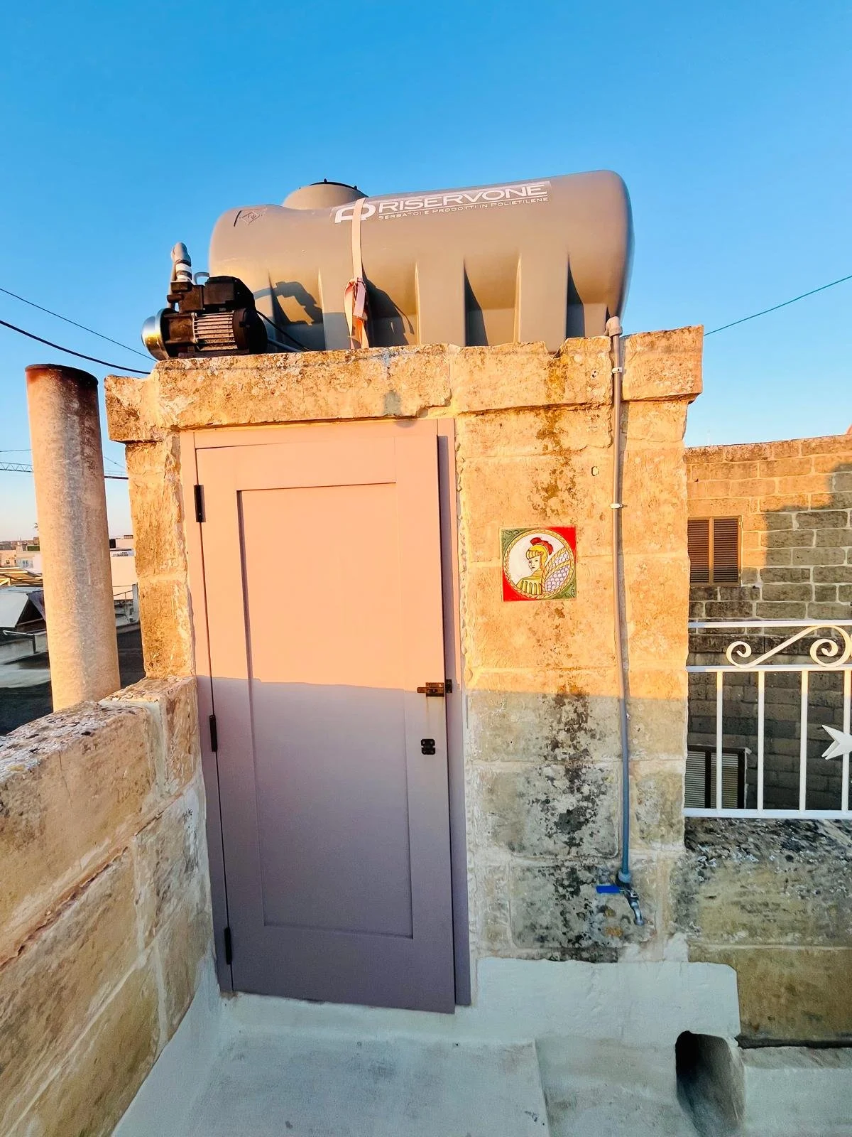 A rooftop structure with a large water tank labeled 'RESERVONE' and a small electric motor, against a clear blue sky. The structure includes a light purple door, stone walls, and a painted decorative tile of a woman in traditional attire. An electric