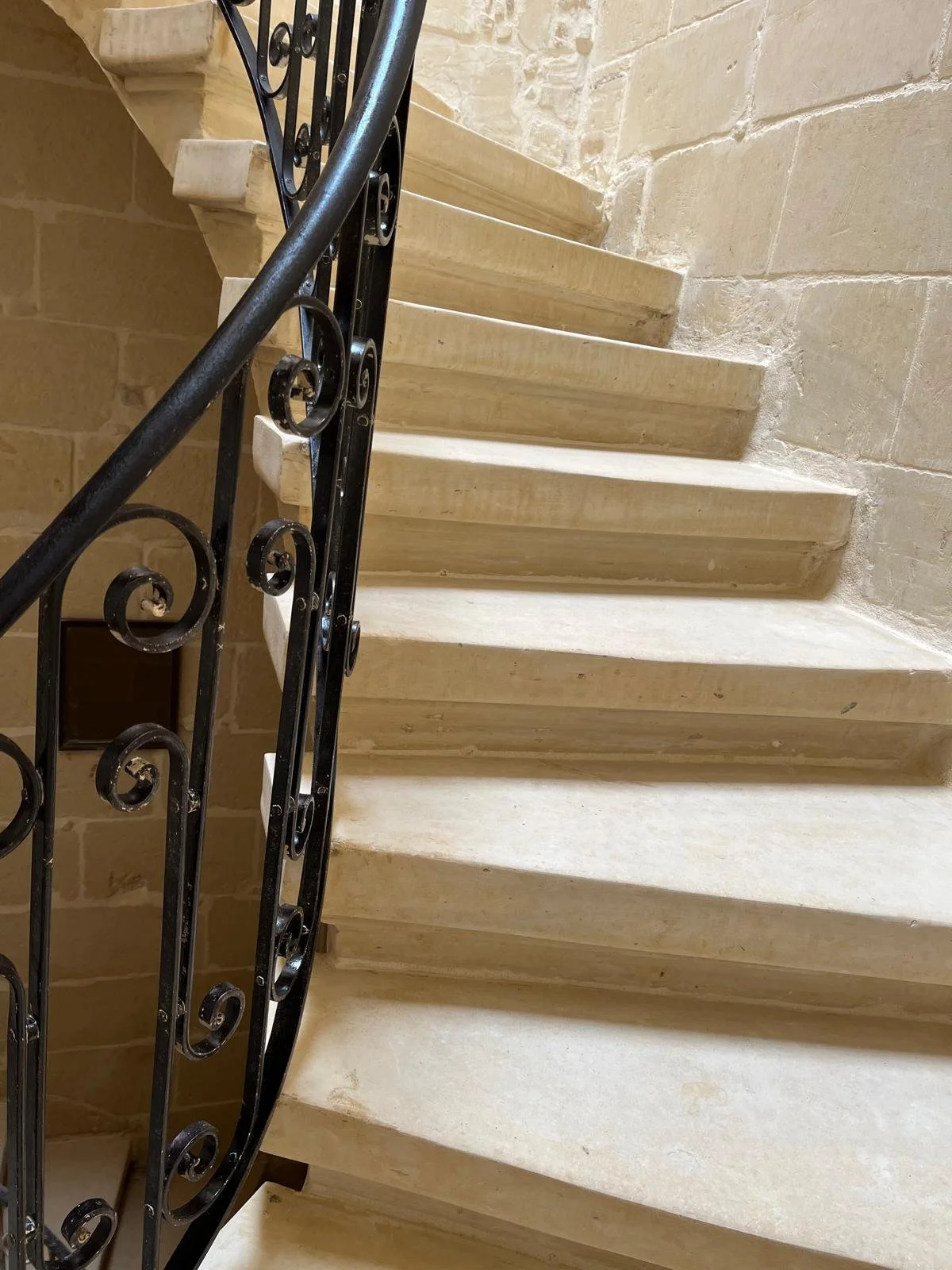 A spiral staircase with beige steps and a black wrought iron handrail against a beige brick wall.