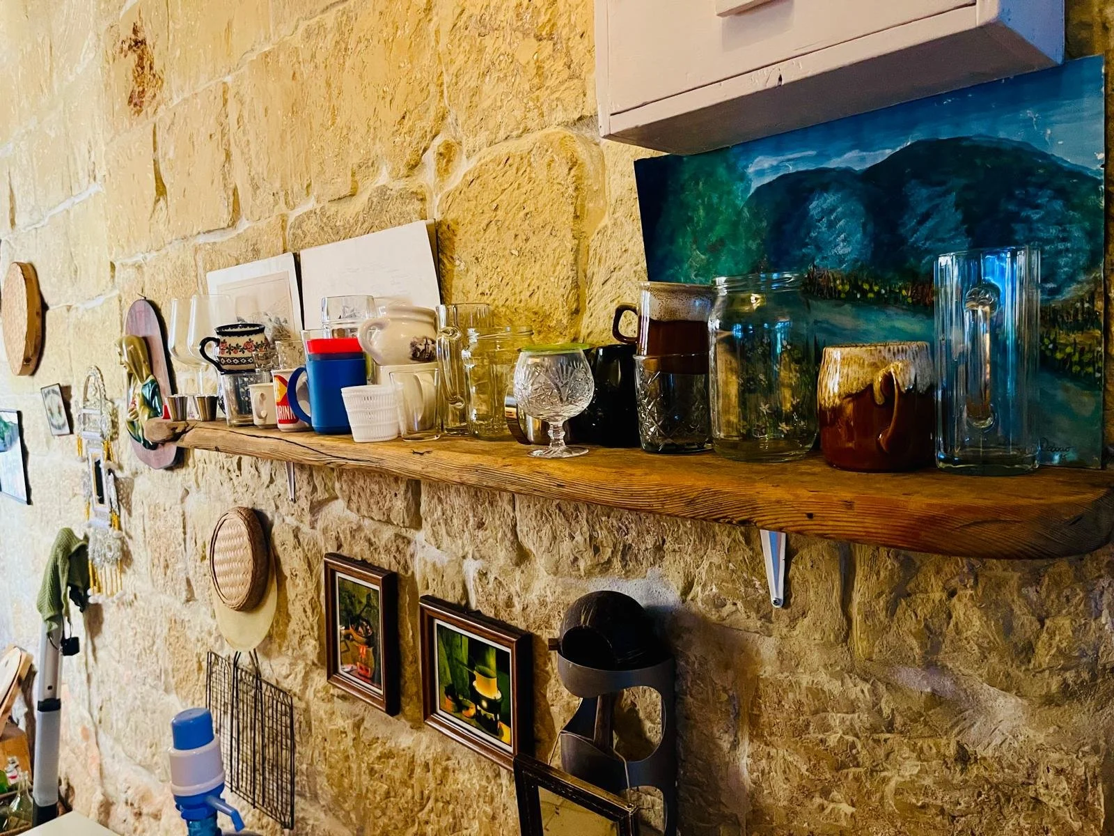 Shelf with various cups, mugs, glasses, and artwork against a stone wall.