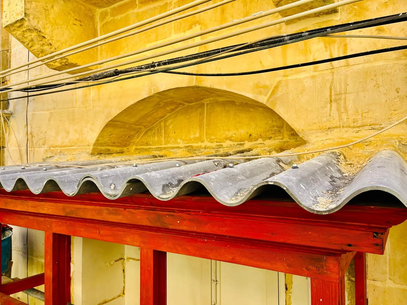 Close-up of a roof with wavy gray tiles on a wooden red-painted structure, with a yellow stone wall and electrical wires in the background.
