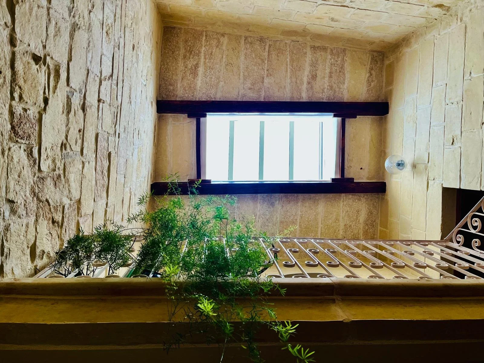 View of a rustic stone interior with a window framed by dark wood beams, a metal railing with decorative curls, a hanging lightbulb, and green plants.