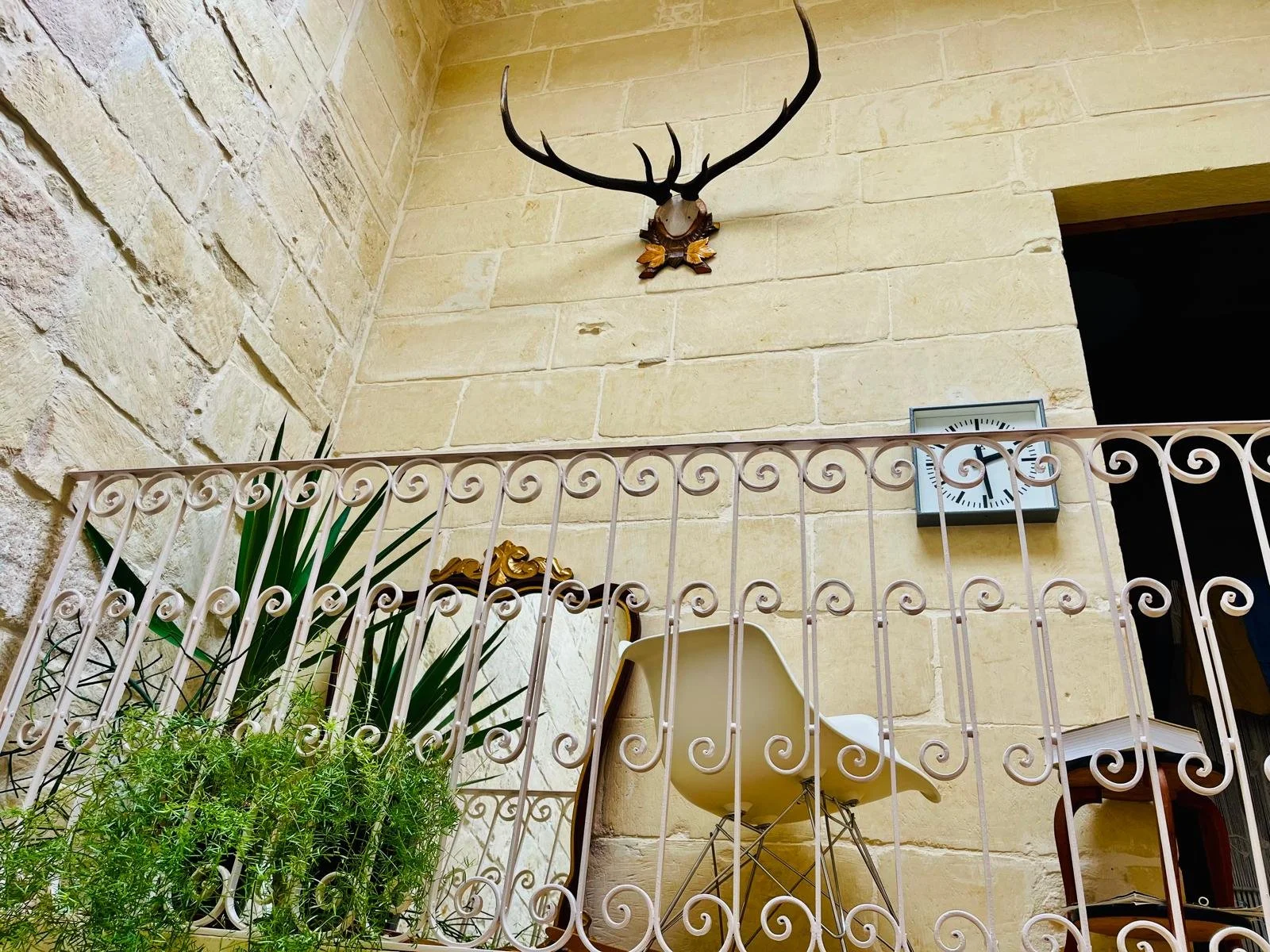 Interior of a balcony with a beige brick wall, a decorative antler mounted on the wall, a clock showing 11:55, green plants, a white chair, and a mirror with a decorative frame.