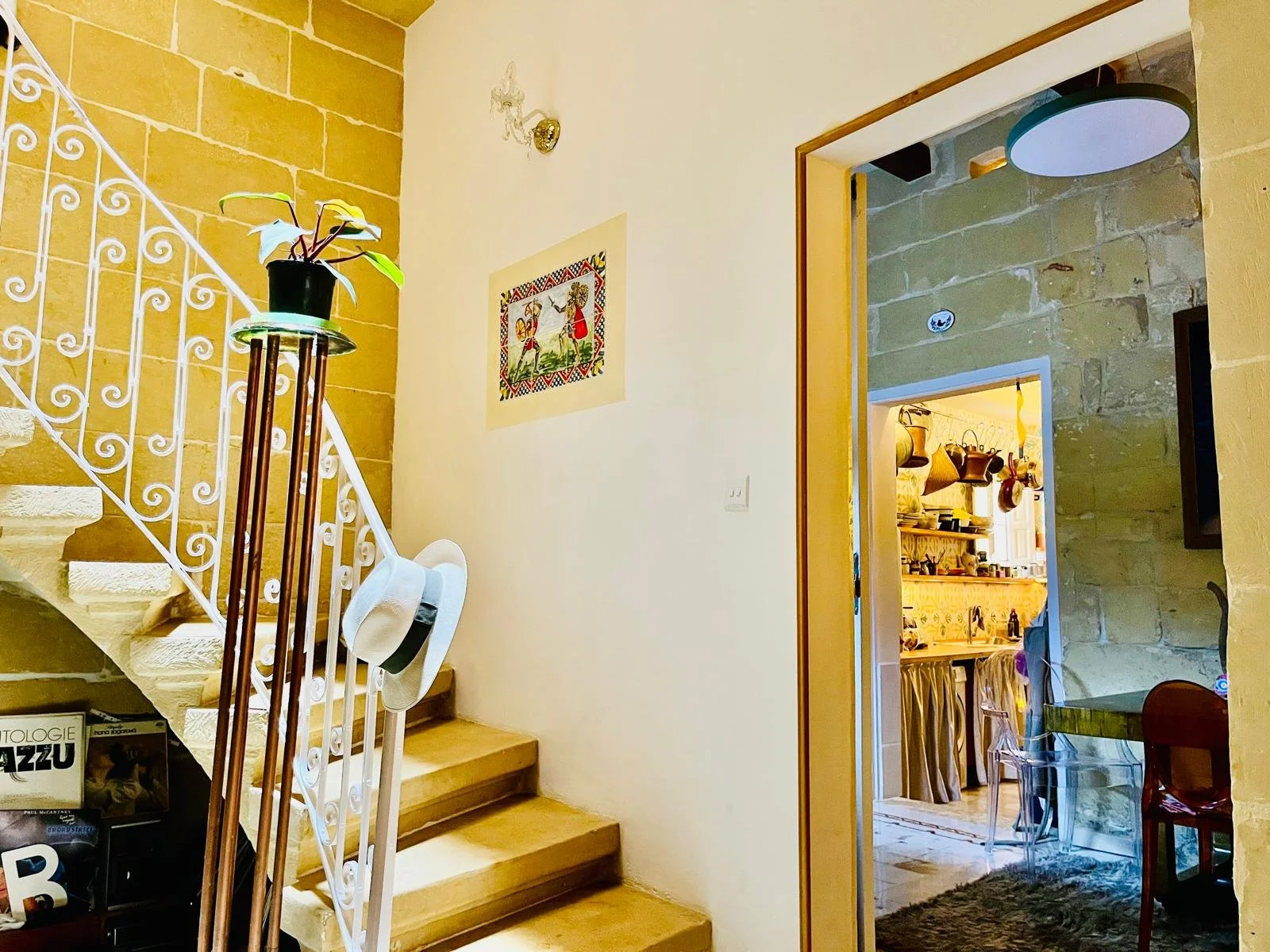 Interior view of a home, showing a staircase with a white wrought iron railing, a hat hanging on the railing, a plant on a tall stand at the base of the stairs, a framed colorful artwork on the wall, and an adjacent room with stone walls, a dining ta