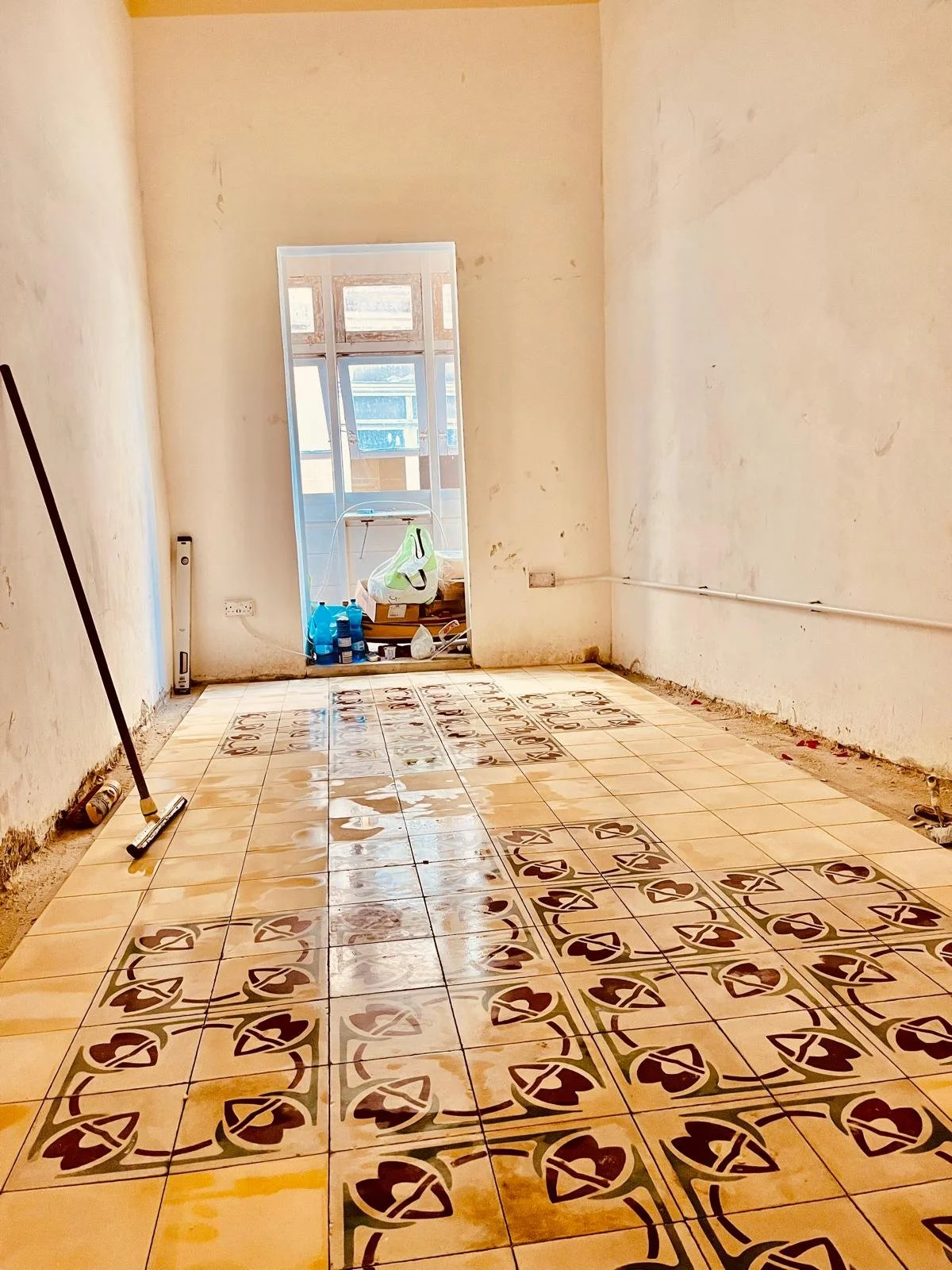 Room under renovation with patterned ceramic floor tiles, a broom leaning against the wall, and supplies near a door leading to a balcony with windows.