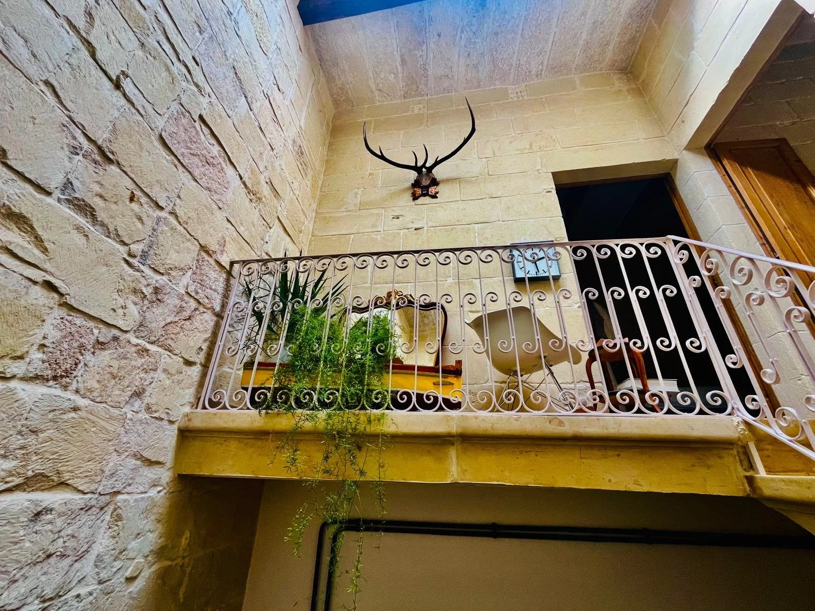 Interior view of a rustic stone and beige brick wall with a decorative metal railing on a balcony. Behind the railing, there are potted plants, a white pottery basin, and a small mirror. An antler-mounted animal head decorates the upper part of the w