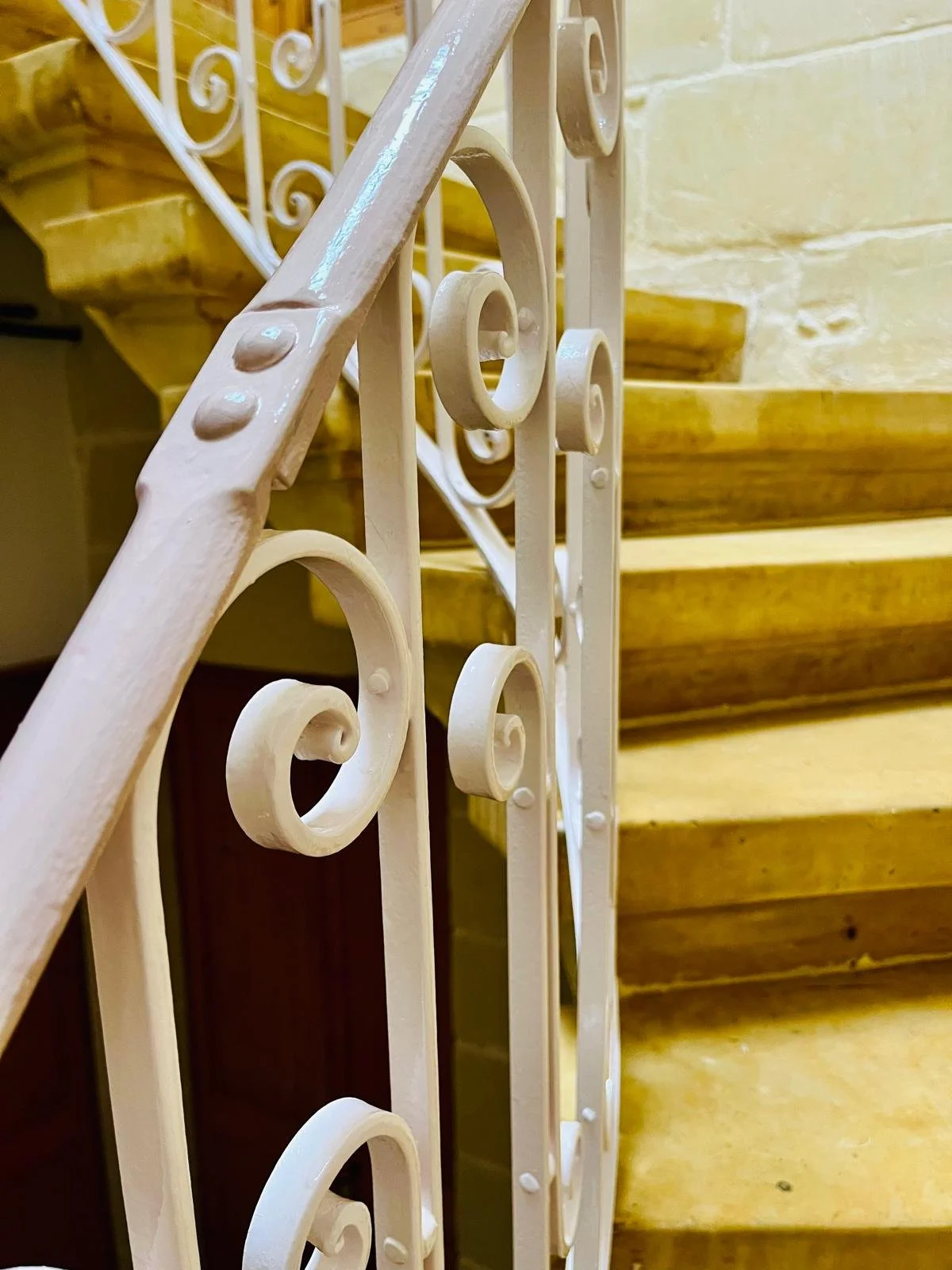 Close-up of a beige decorative metal railing on a staircase, with wooden stairs in the background.