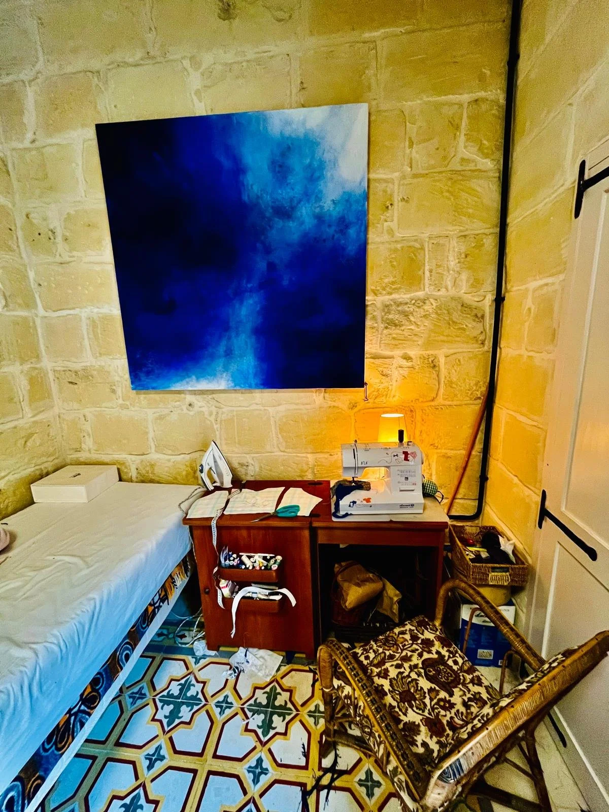 A sewing workspace with a bed, a small wooden table, and a wicker chair. The table has a sewing machine and a lamp. The wall has a large blue abstract painting, and the floor has a colorful geometric tile pattern.