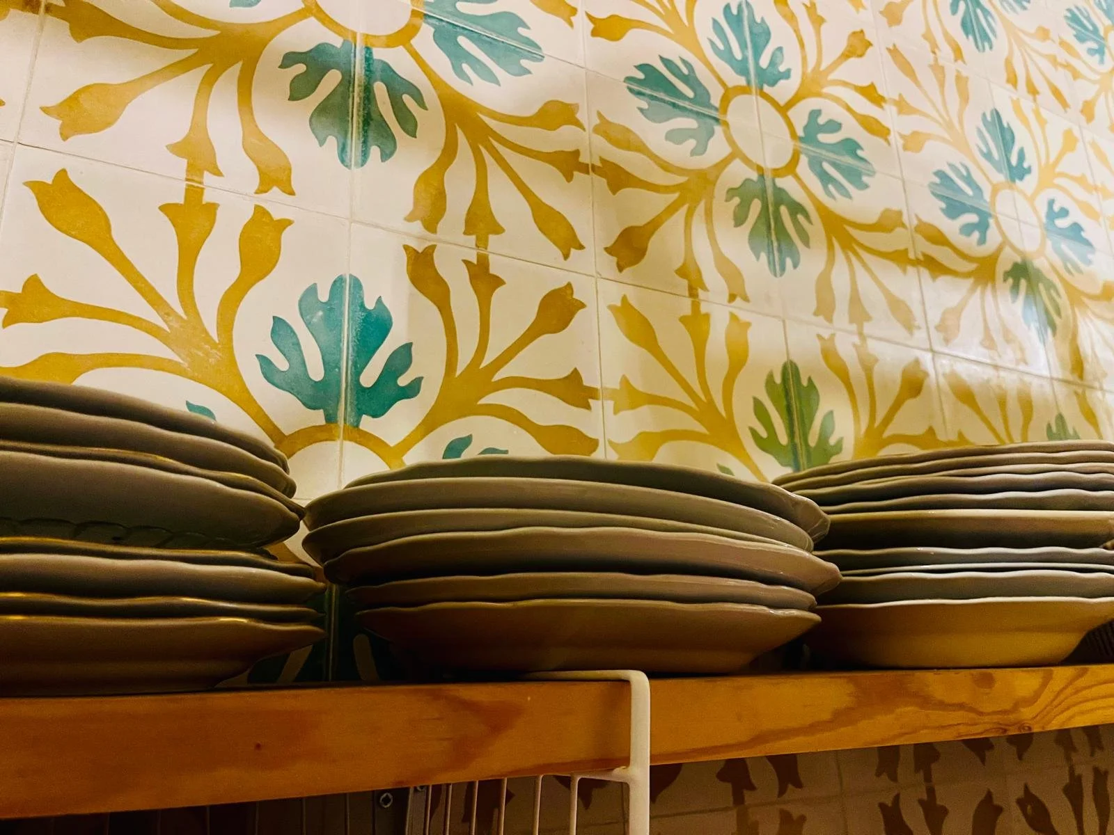 Stacks of white plates on a wooden shelf against a colorful tiled wall with yellow, green, and blue floral patterns.