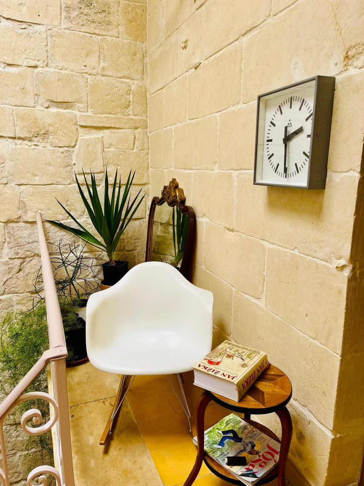 A cozy corner with a brick wall, a white modern chair, a small wooden side table holding a large book, a smaller magazine underneath, a potted plant, a vintage mirror, and a wall clock showing approximately 4:05.