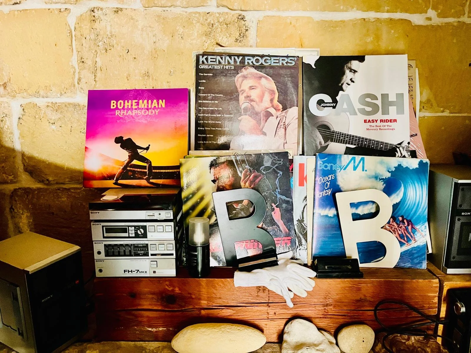 A collection of vinyl records, magazine, and electronic music equipment on a wooden table, set against a stone and brick wall. The records include Bohemian Rhapsody, Kenny Rogers, Johnny Cash, and Boney M.