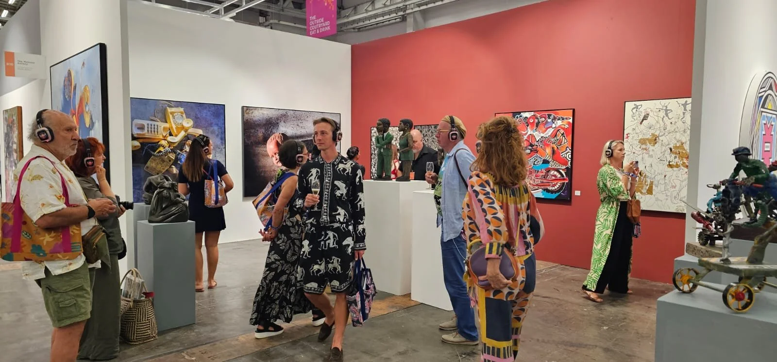 People at an art gallery viewing paintings and sculptures, some with glasses of wine.