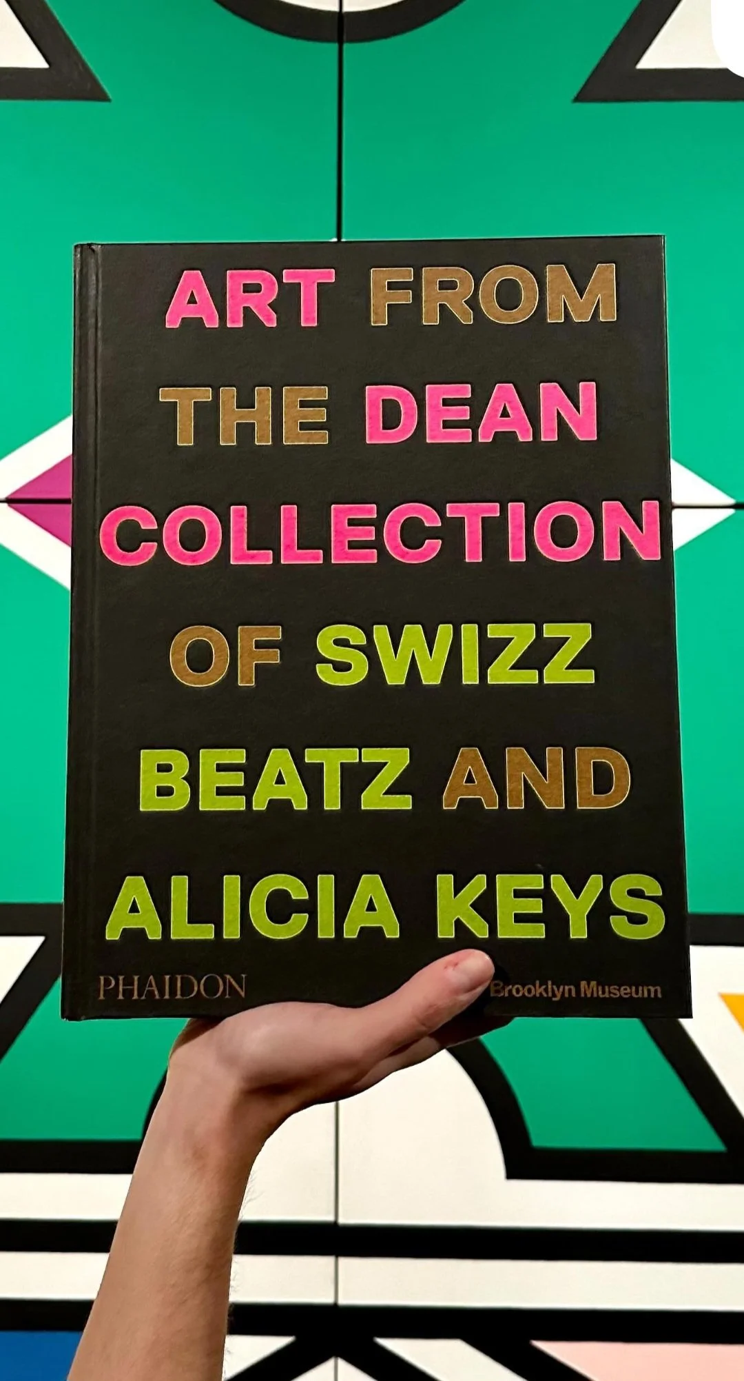 Hand holding a book titled 'Art from the Dean Collection of Swizz Beatz and Alicia Keys' in front of a colorful geometric background.