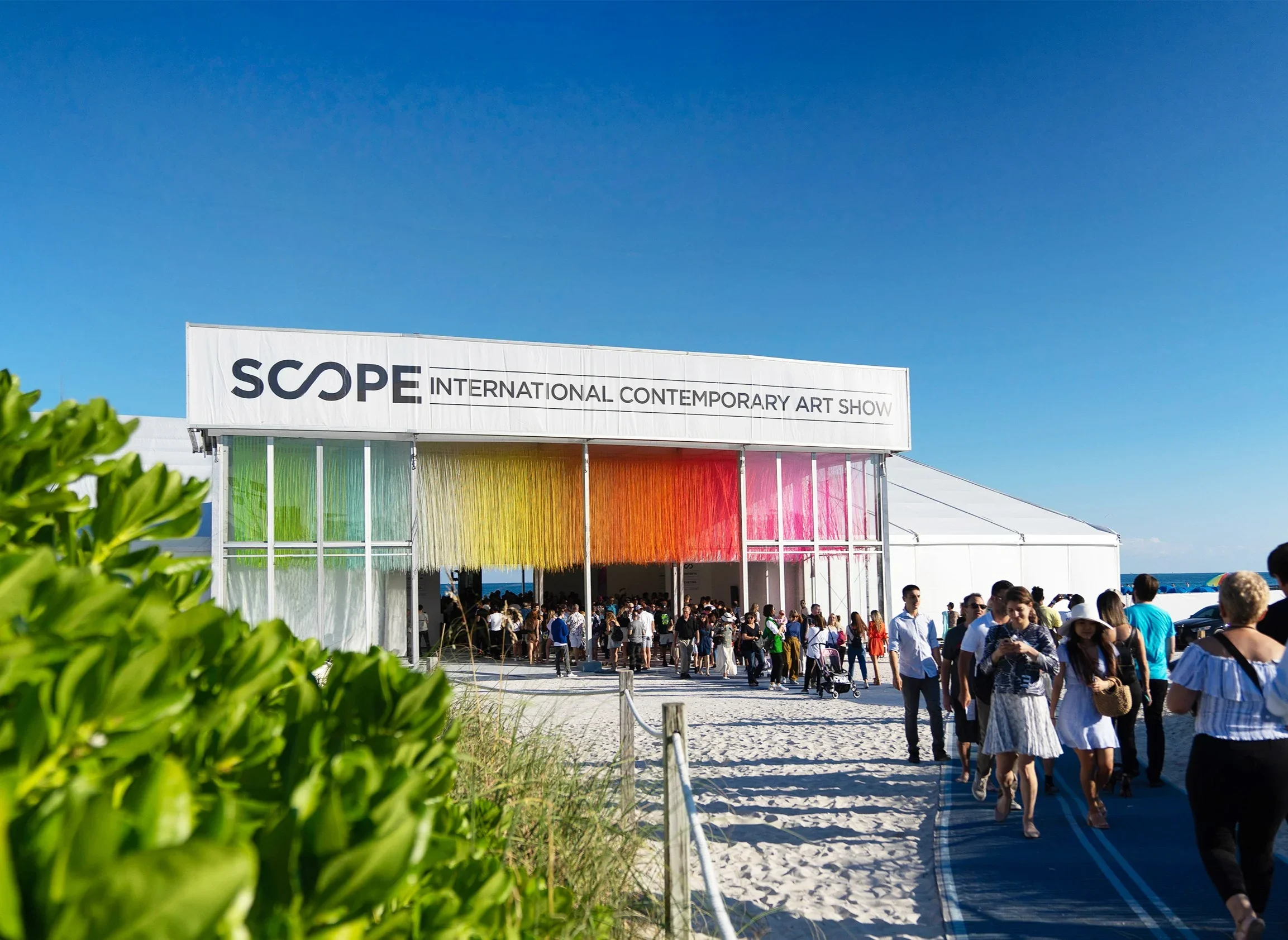 People entering the Scope International Contemporary Art Show in a large white tent on a beach with bright blue sky, featuring terrazzo flooring, green shrubbery in foreground, and colorful hanging decorations inside the tent.