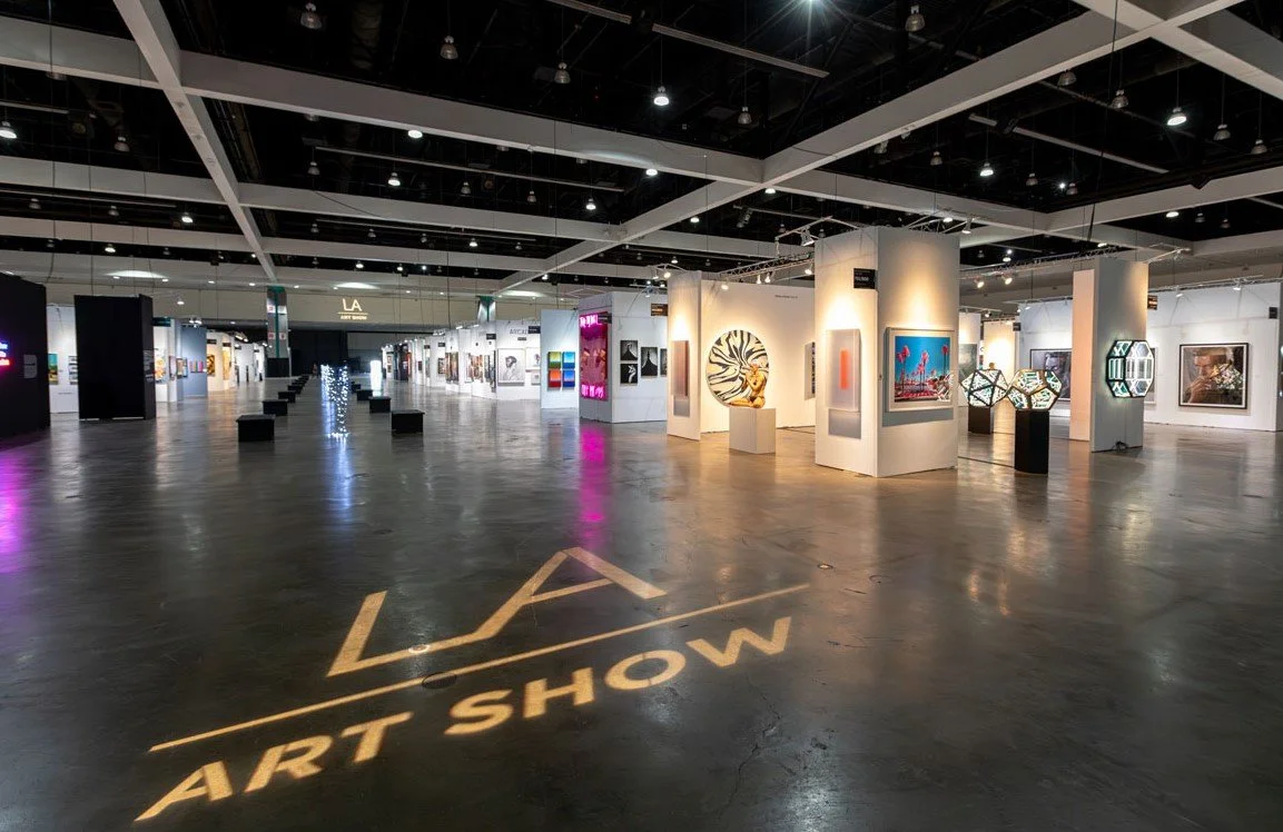 Interior of a modern art gallery with various artworks displayed on white walls, and a sign on the floor reads "LA ART SHOW."