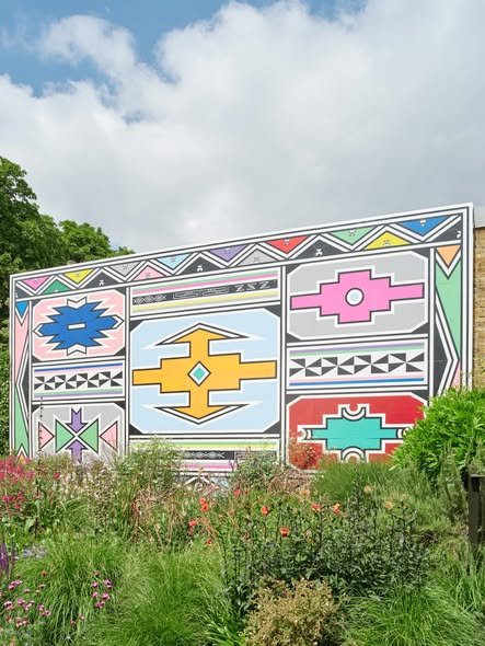 Large outdoor artwork featuring geometric patterns and shapes in bright colors including pink, blue, yellow, green, red, and purple, set against a partly cloudy sky with greenery in the foreground.