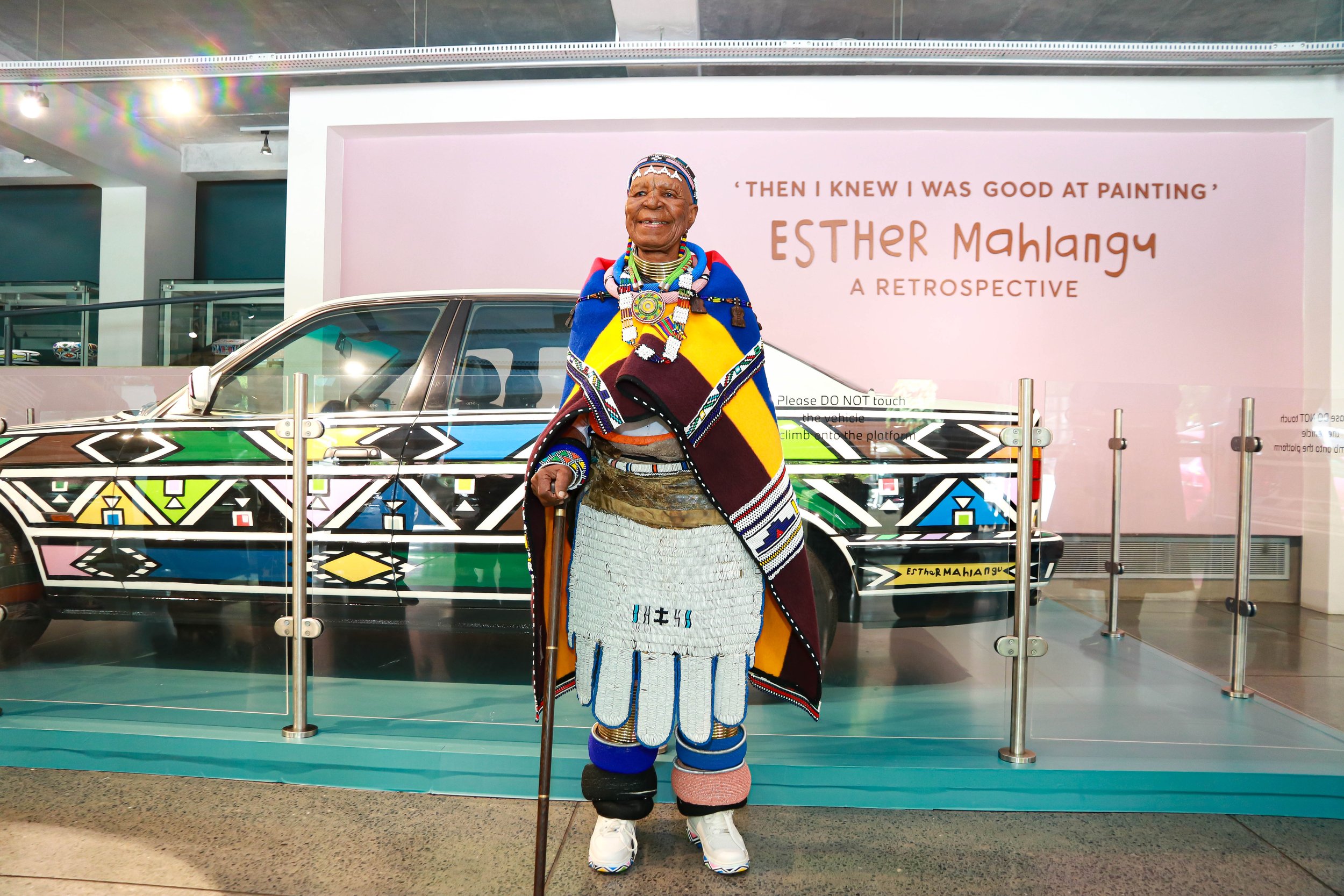 An elderly woman in traditional Maasai attire standing inside a museum, in front of a colorful geometric-patterned vehicle, with a pink sign that reads "Then I knew I was good at painting" Esther Mahlangu, a retrospective.