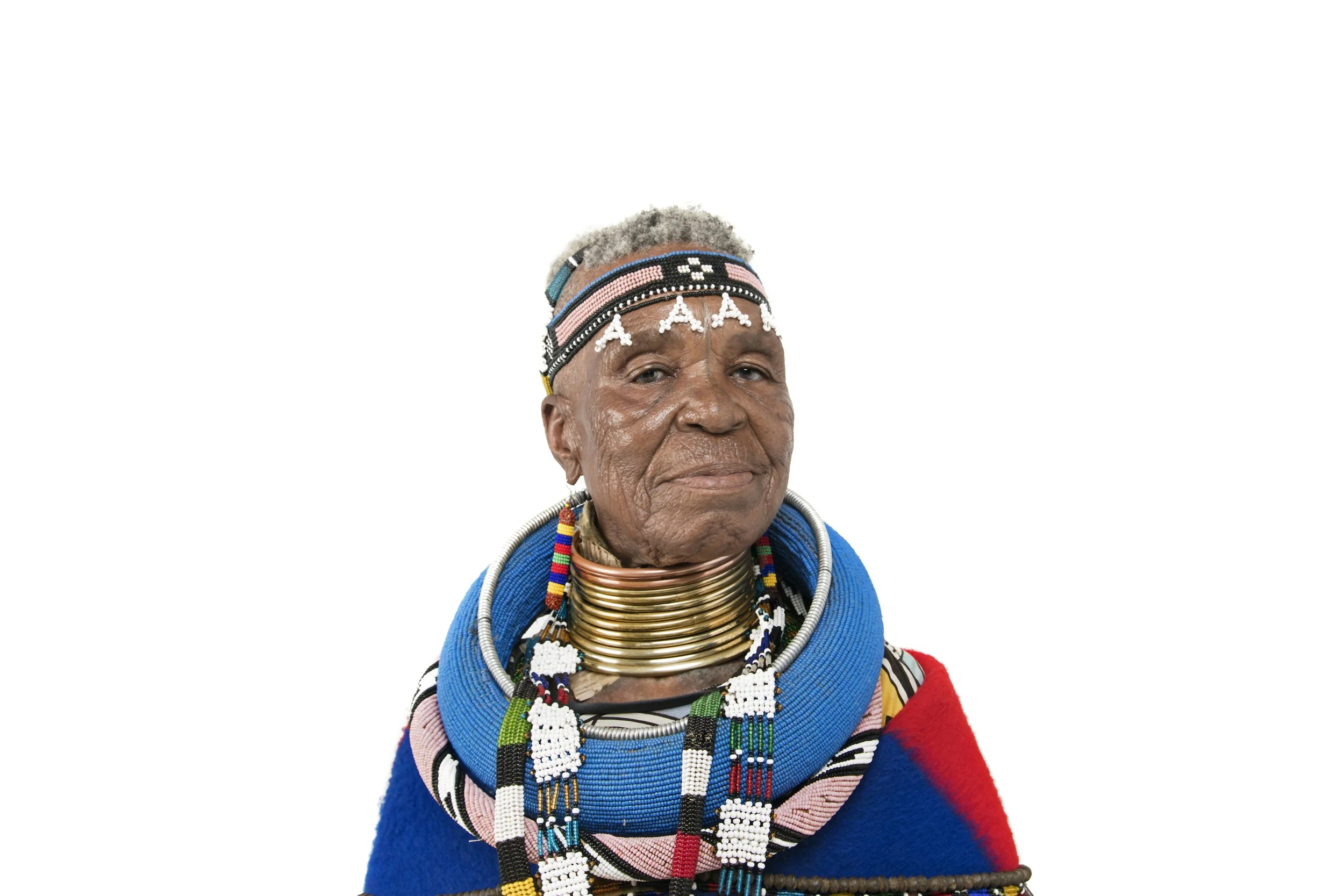 An elderly African woman dressed in traditional Maasai attire and jewelry, with layered beaded necklaces, earrings, a colorful shawl, and beaded headband with the letters 'AAA' written on it.
