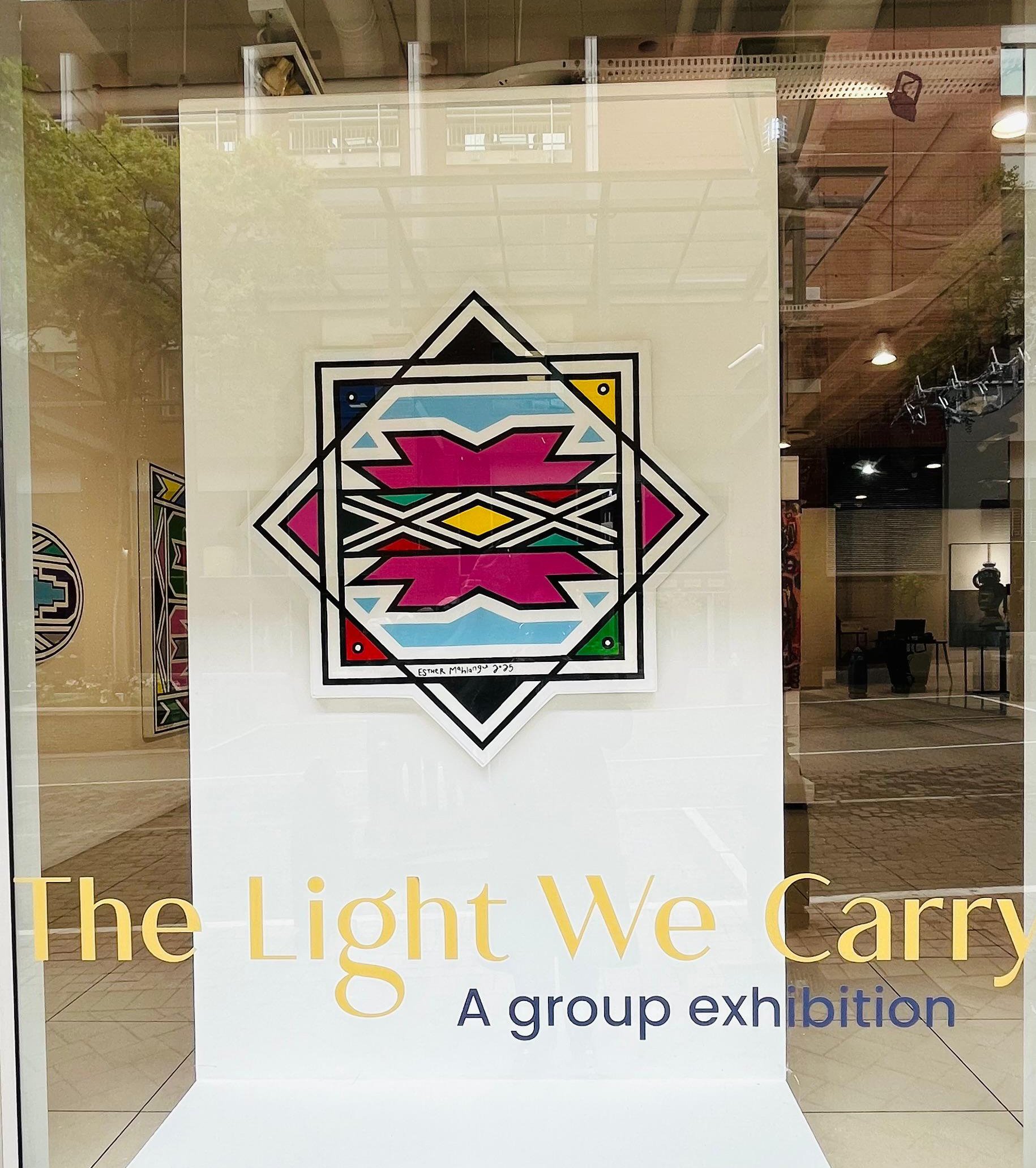 A colorful geometric art piece with symmetrical patterns in pink, yellow, green, blue, black, and white, displayed in a gallery window with the text 'The Light We Carry: A group exhibition' on the glass.