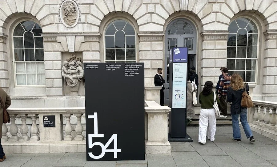 People standing outside a museum or building with large arched windows and ornate stone decorations, including a lion sculpture. There are information signs and a ticket or entrance counter.
