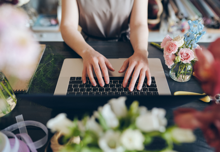 Person working on a laptop surrounded by flowers and craft supplies.