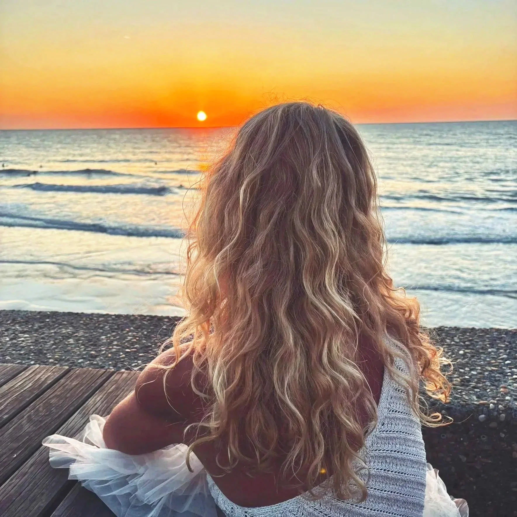 Une jeune fille aux cheveux bouclés assise sur un quai en regardant un coucher de soleil sur la mer.