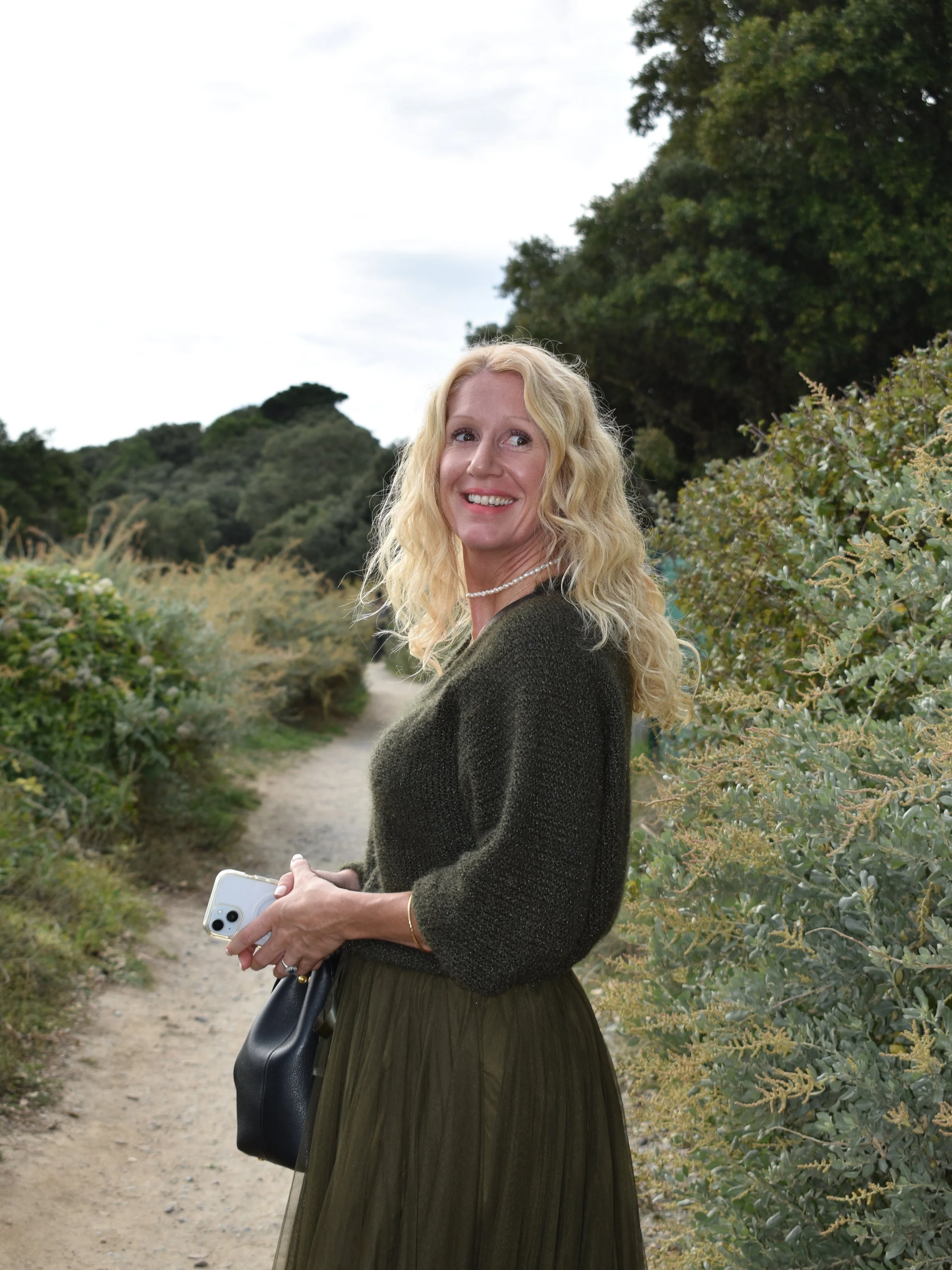 Femme blonde souriante marchant sur un sentier dans la nature avec des arbres et des buissons environnants, tenant un téléphone et un sac à main.