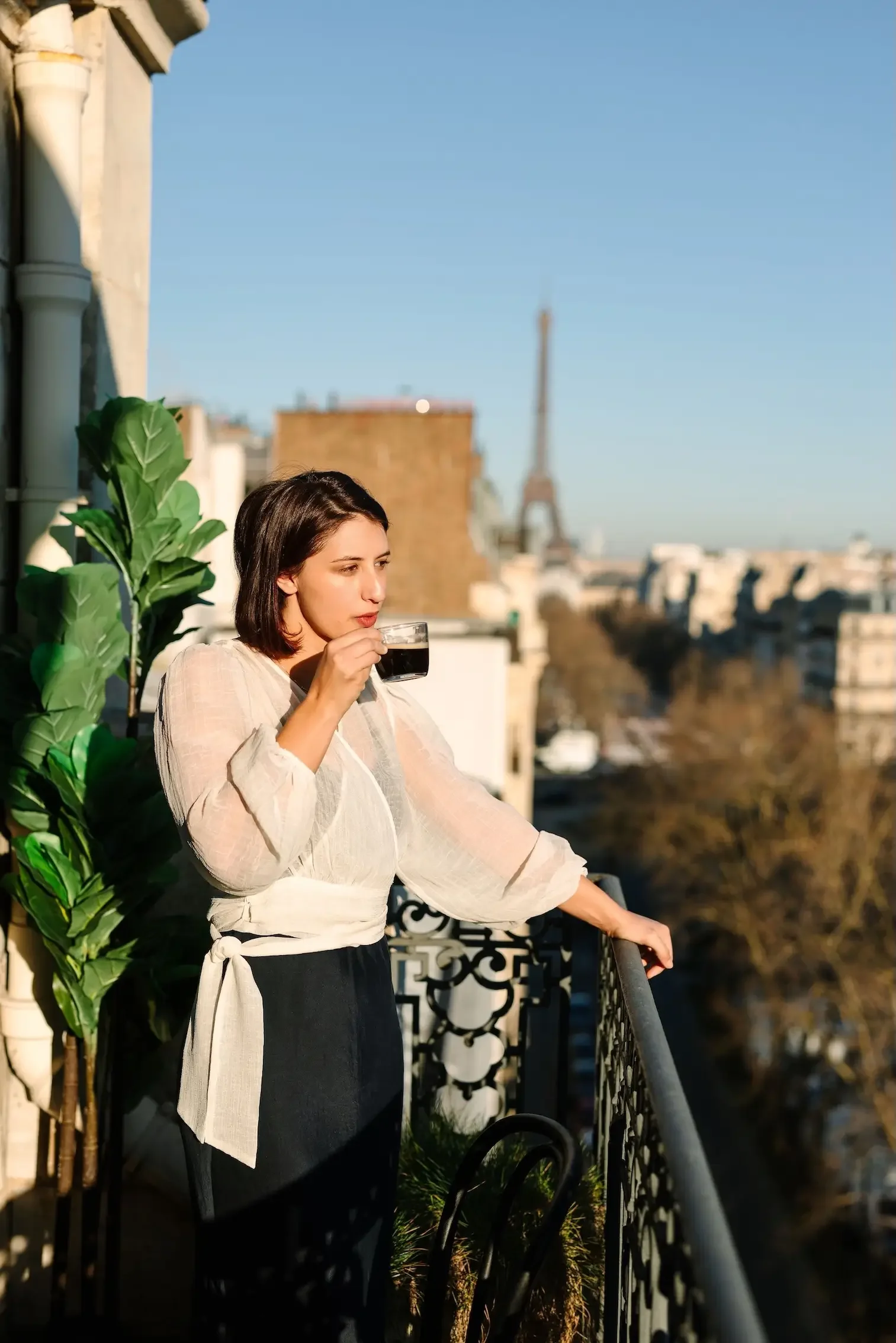 Hélène with dark hair drinking coffee on a Parisian balcony with the Eiffel Tower in the background during a sunny day.