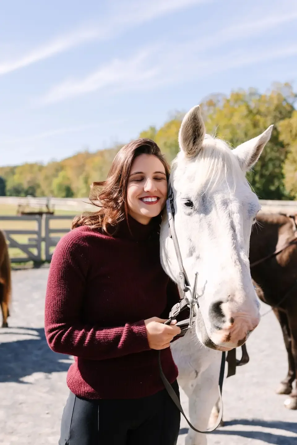 Hélène with brown hair smiling and hugging a white horse outdoors on a sunny day with trees in the background.