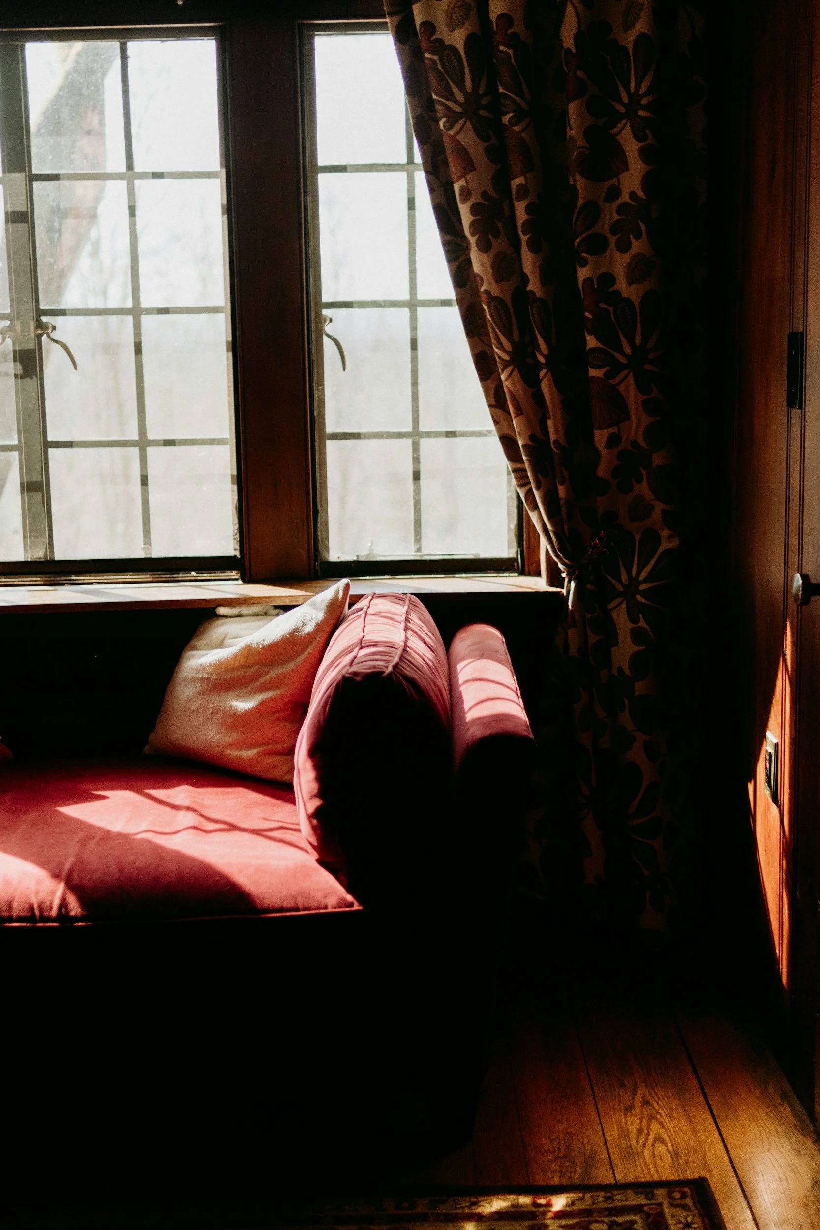Sunlight streaming through a window illuminating a cozy armchair with red upholstery, a white pillow, and floral curtains in a warm-toned living room.