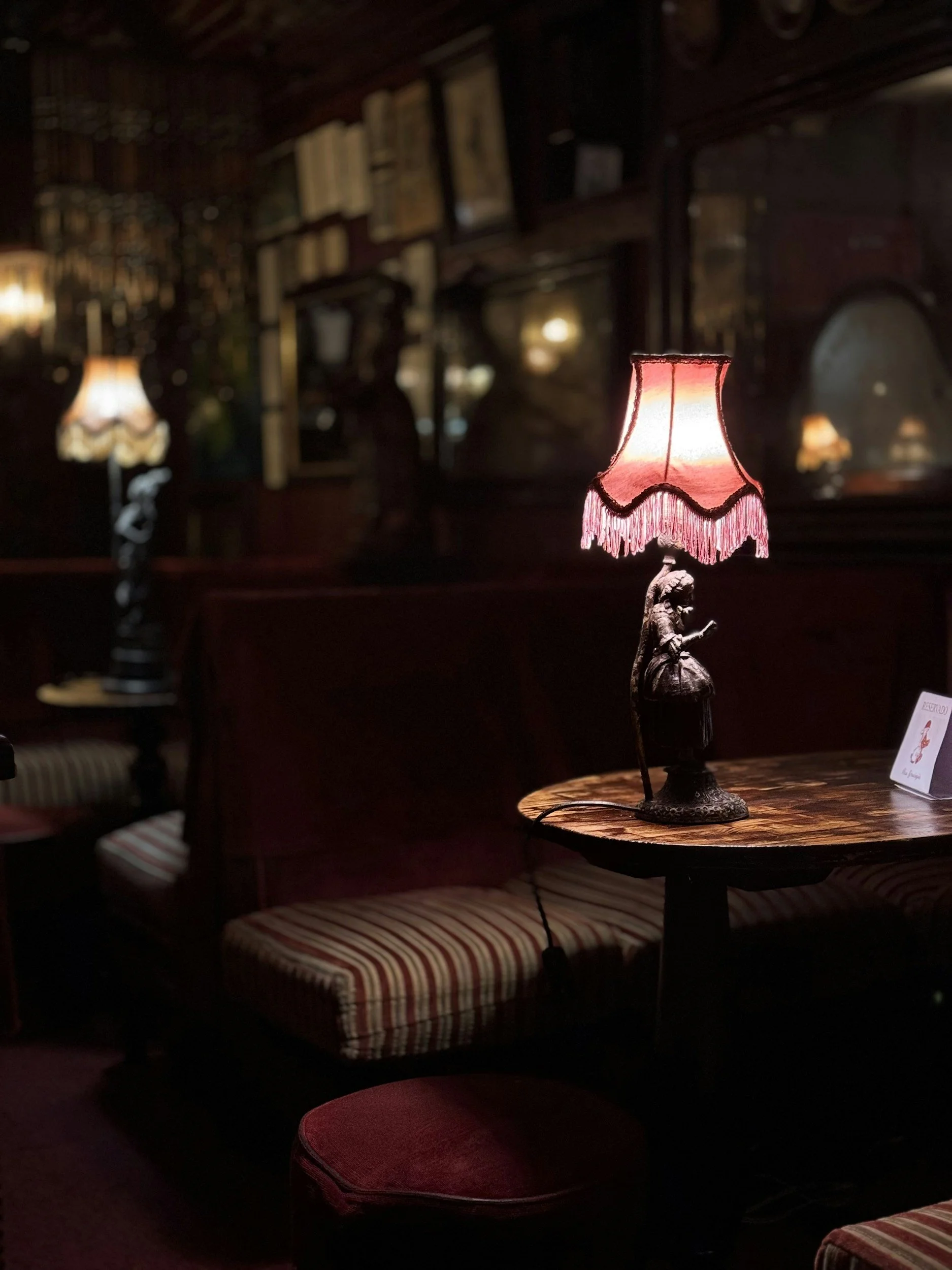 Dimly lit vintage-style restaurant or lounge with a small table illuminated by a pink lamp featuring a figurine of a woman reading a book and dressed in period clothing.
