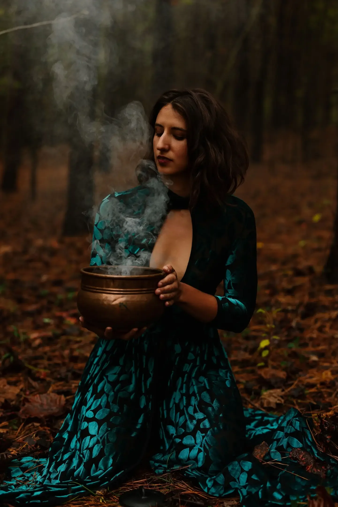 Hélène with dark hair kneeling in a forest, holding a steaming bowl, dressed in a dark blue and teal patterned dress.