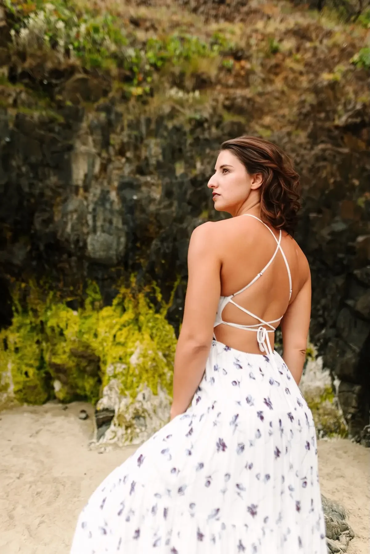 Hélène with short brown hair standing outdoors on sandy ground near a rocky cliff with green foliage in the background, wearing a white dress with a floral pattern and a backless top with crisscrossing straps.