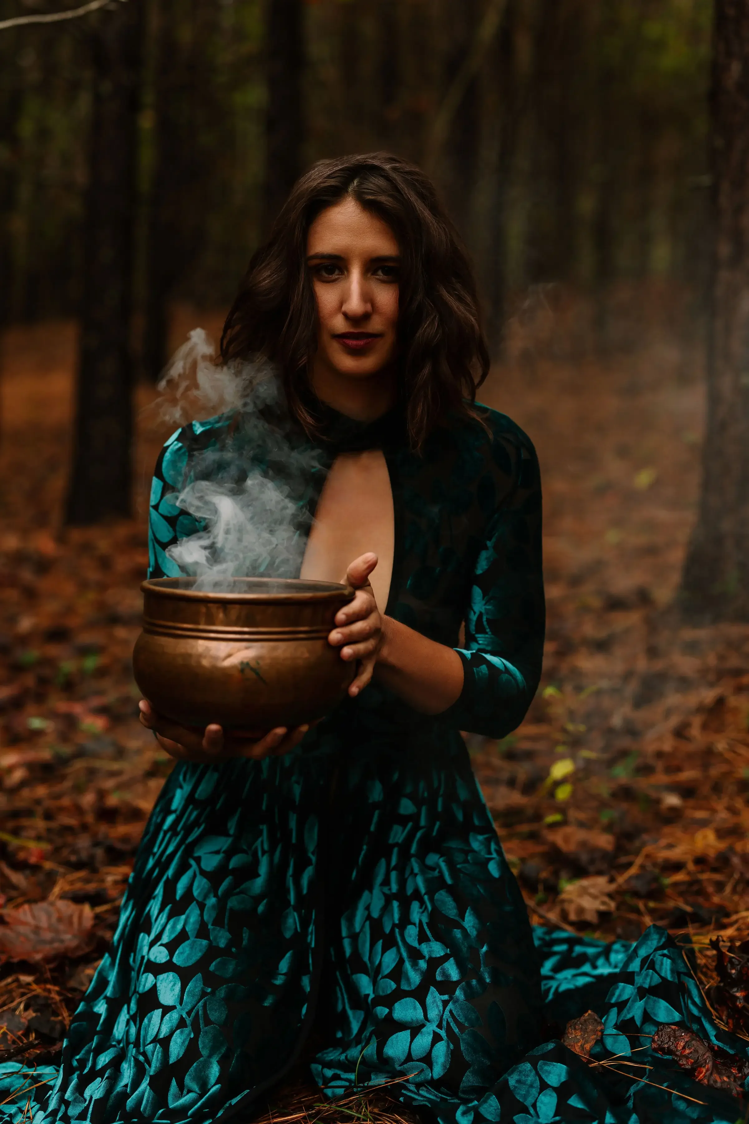 Hélène with dark wavy hair sitting on the forest floor holding a steaming wooden bowl, surrounded by autumn leaves and trees.