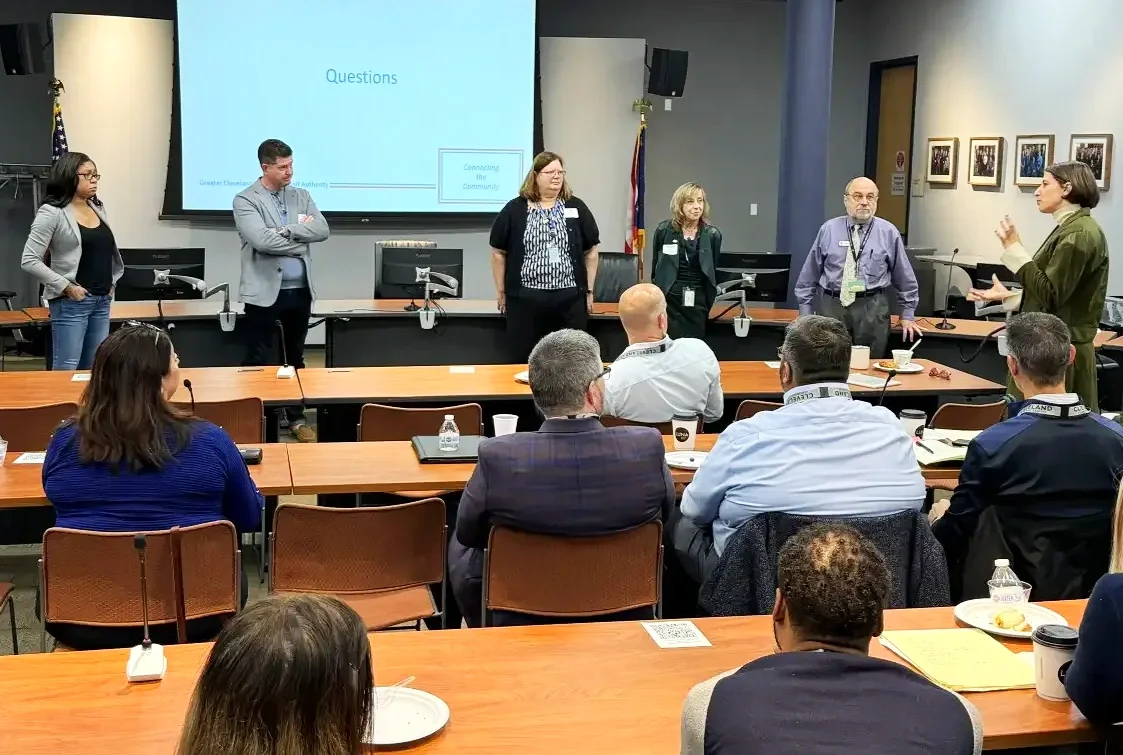 Panel discussion with five people standing at the front of a conference room, audience seated and listening, large screen displaying 'Questions', flags, and framed photographs on the wall.