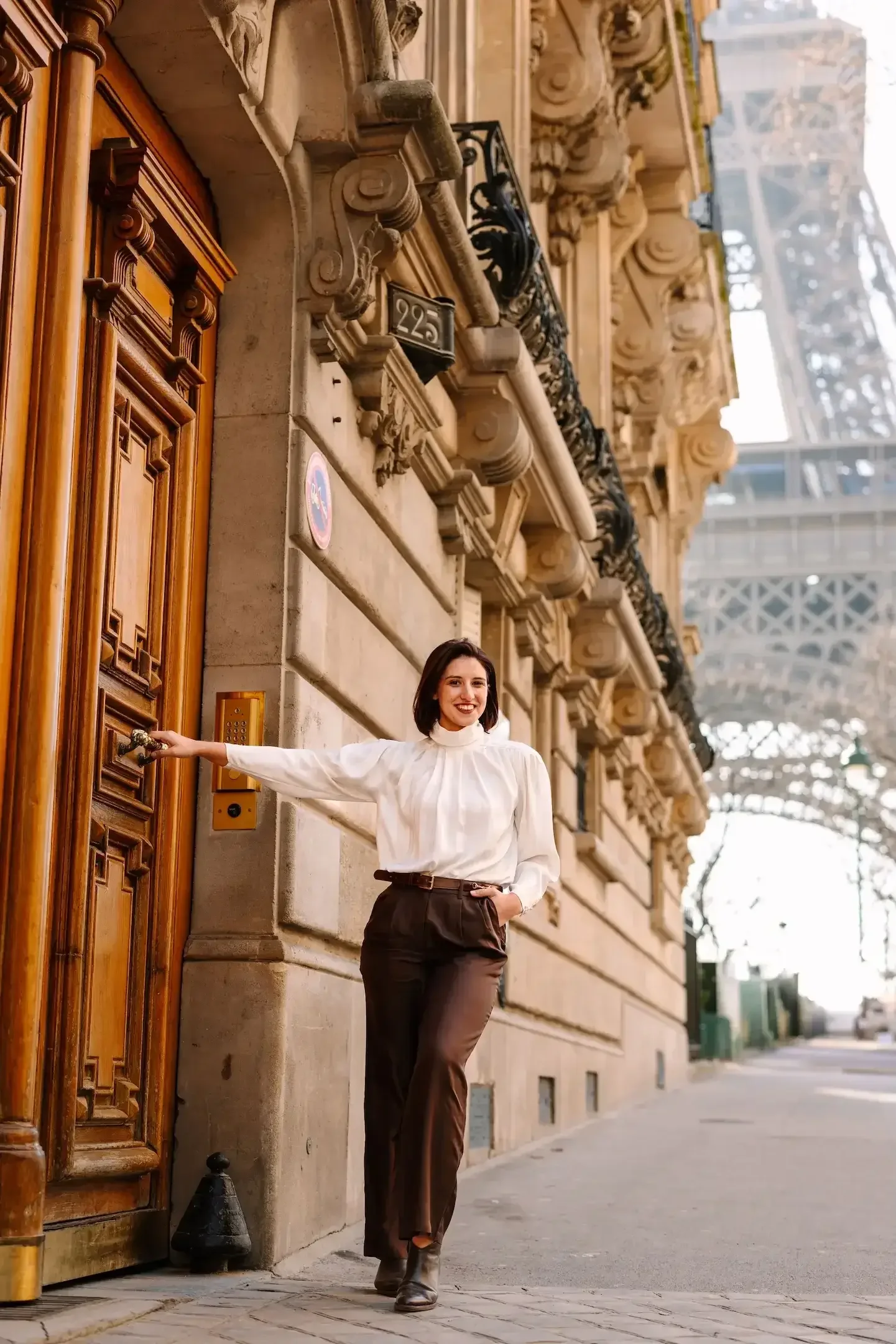 Hélène smiles while reaching for a doorbell on a historic stone building with ornate architecture and a wooden door in Paris, France with the Eiffel Tower in the background.