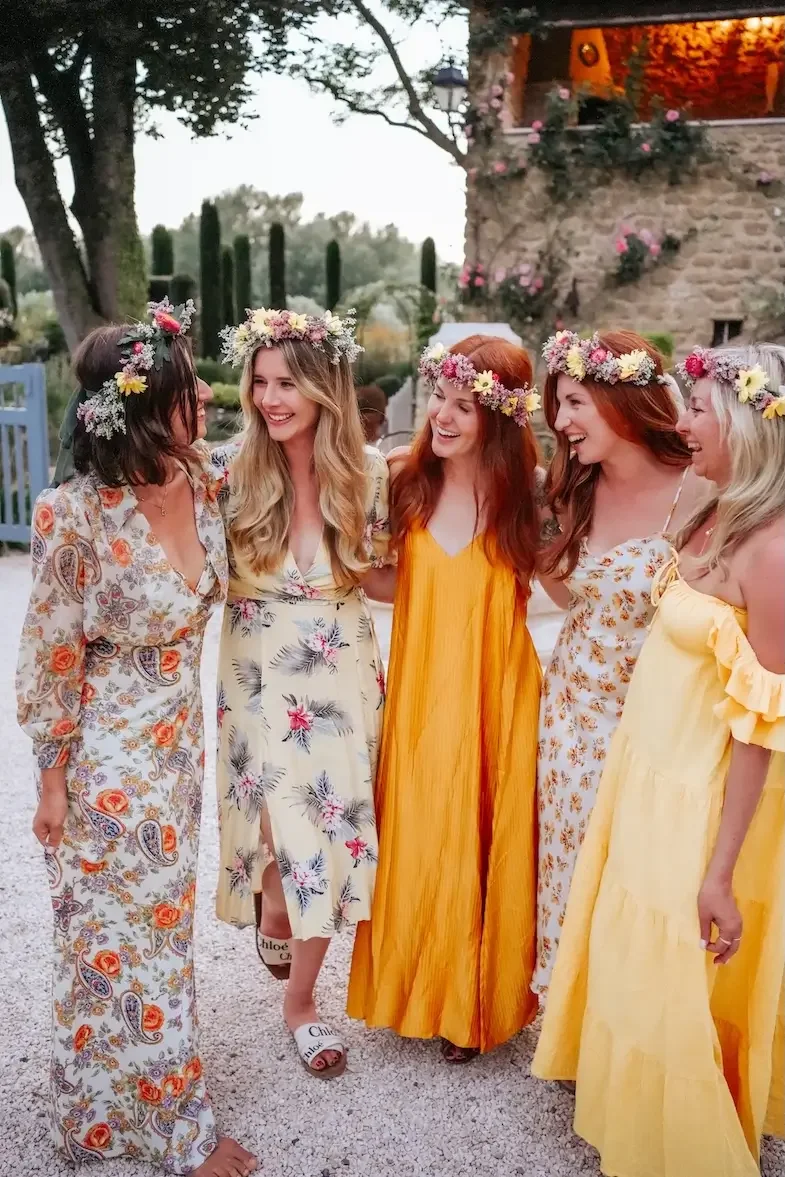 Group of six women wearing floral dresses and flower crowns, standing outdoors and smiling.