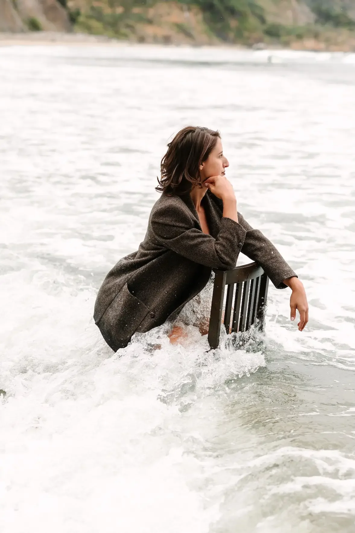 Hélène in a brown blazer sitting on a chair in the water, with her chin resting on her hand, looking contemplative.