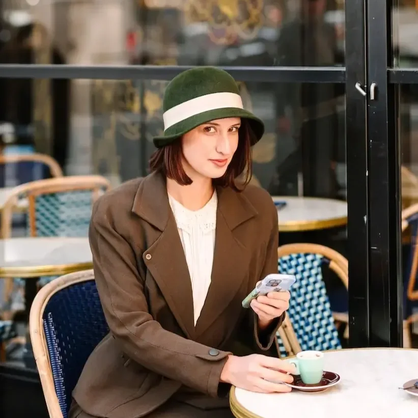 Hélène in a brown blazer and white blouse sitting at an outdoor café in Paris holding a phone in one hand and a small cup of coffee on a saucer with the other. She's wearing a green hat with a white band and looking at the camera with a slight smile.