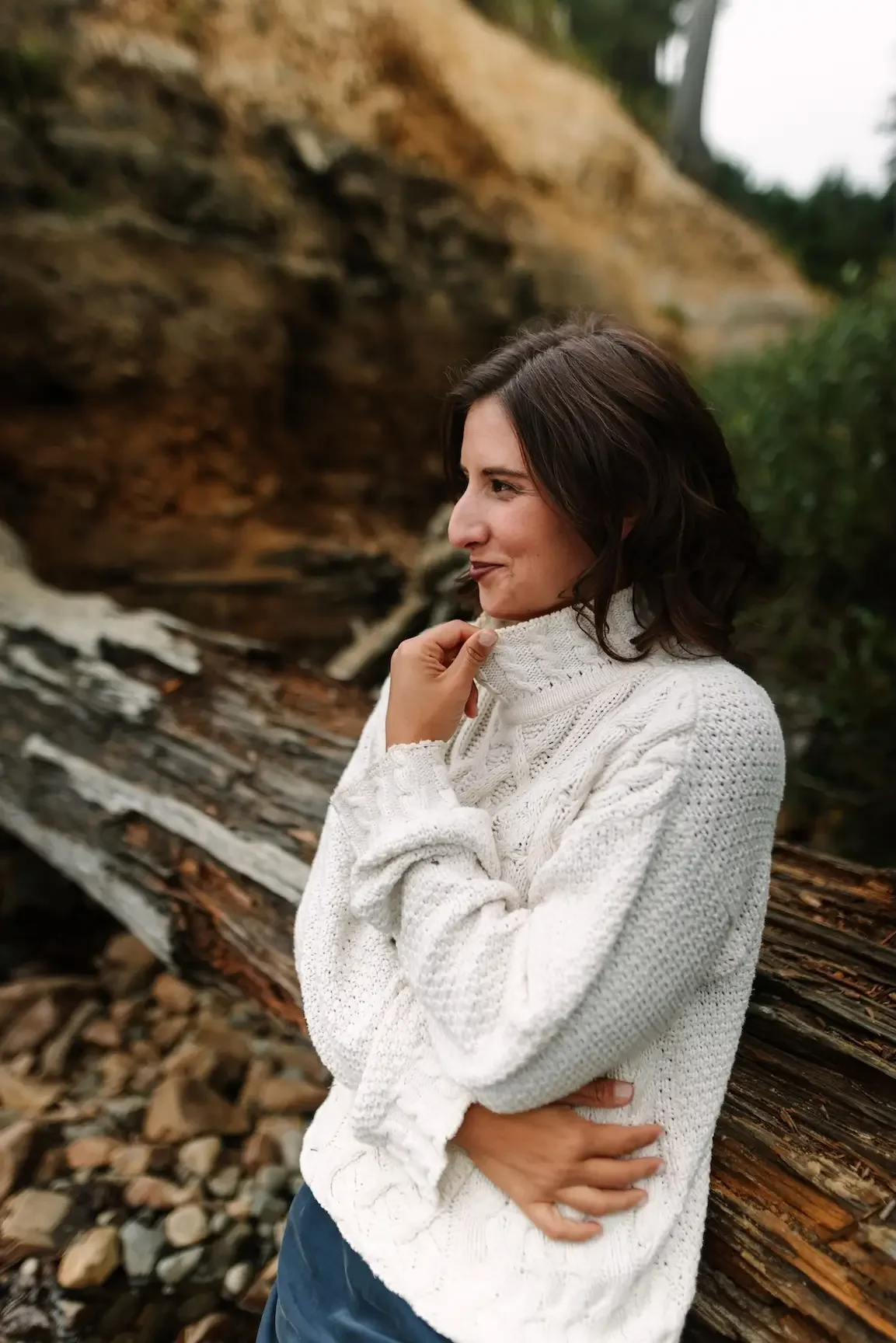 Hélène in a white knit sweater standing outdoors near a fallen tree and rocky terrain, with a hillside and trees in the background.